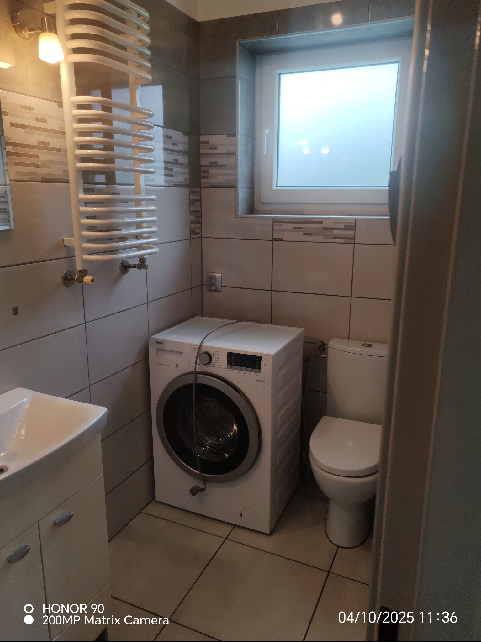 Ustawiona w rogu łazienki pralka Beko, obok toaleta i umywalka z szafką. Na ścianie płytki w odcieniach szarości i biały grzejnik drabinkowy. Okno z białymi ramami.