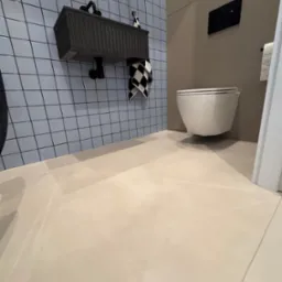 Nowoczesna łazienka z mikrocementową podłogą, wiszącą toaletą i umywalką na tle niebieskich płytek. Minimalistyczny design, gładka powierzchnia podłogi.