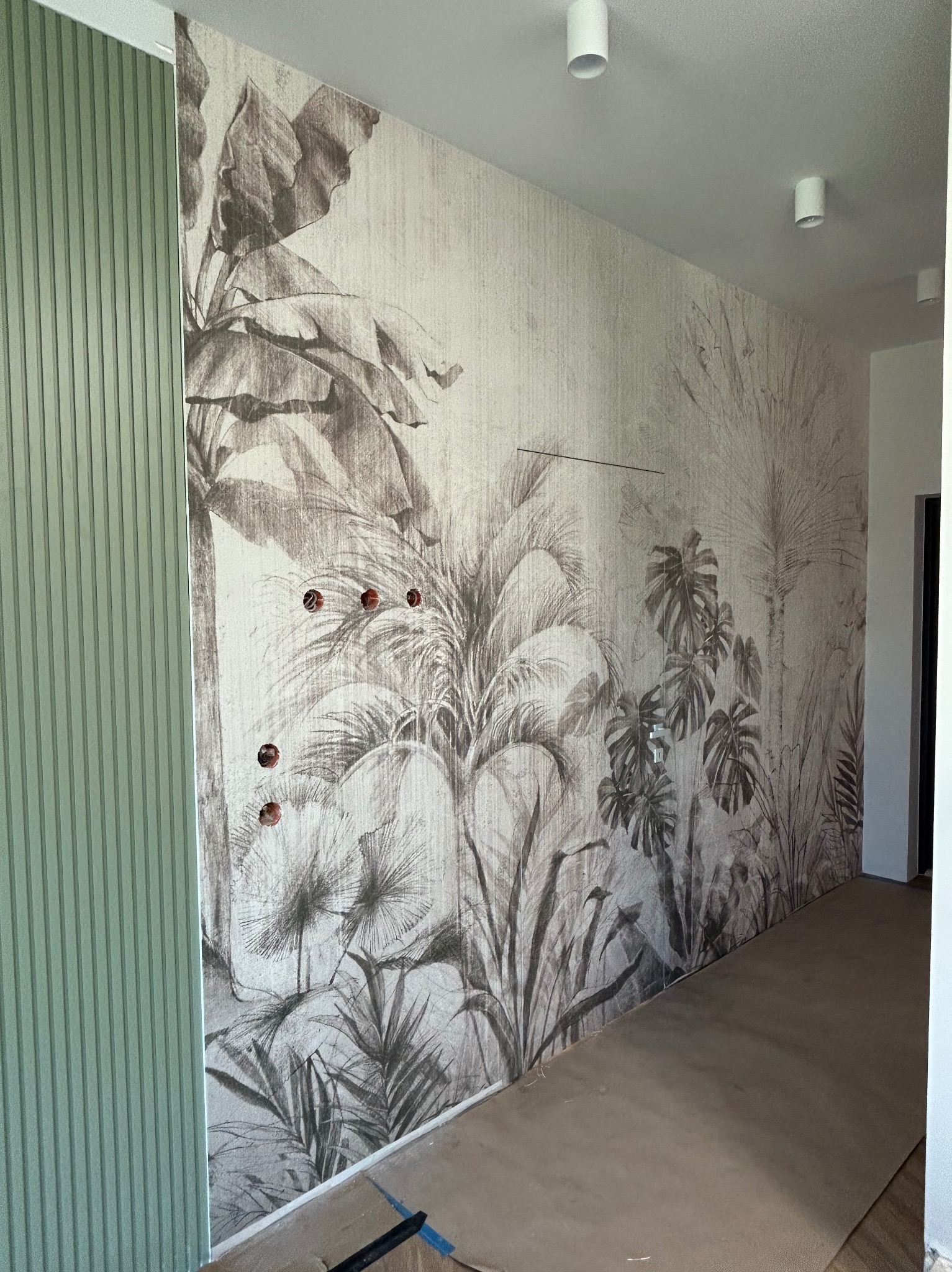 Ściana z fototapetą w motywy roślinne, widoczne puszki elektryczne. Obok ściana z pionowymi panelami w kolorze zielonym. Nowoczesne oświetlenie sufitowe.