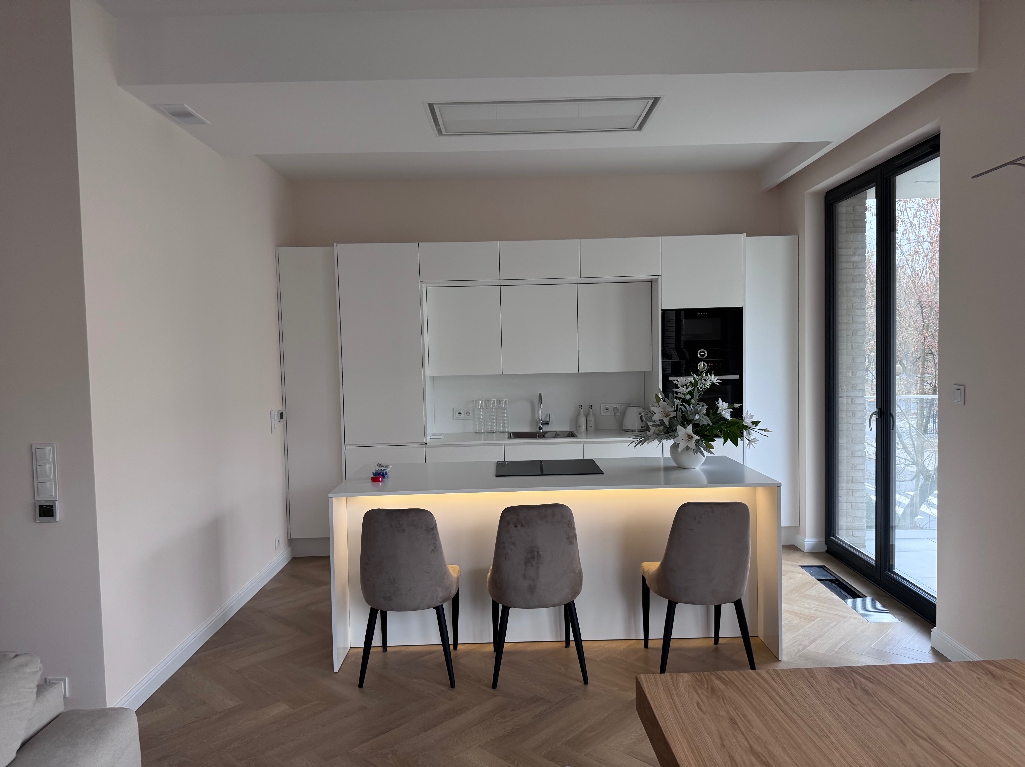 Nowoczesna, jasna kuchnia z wyspą oświetloną od dołu, trzema krzesłami i parkietem w jodełkę. Minimalistyczny design z dużym oknem balkonowym.