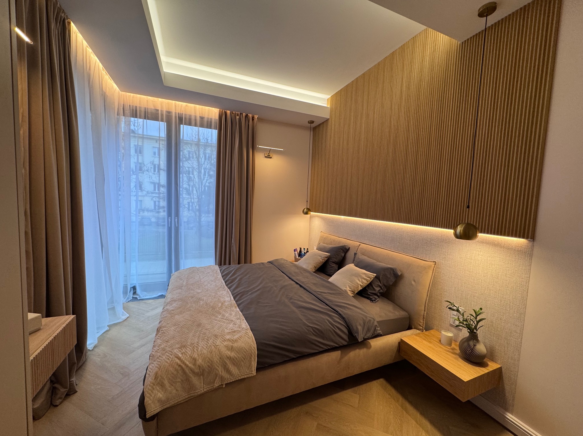 Nowoczesna sypialnia z drewnianą ścianą, podświetlanym zagłówkiem i minimalistyczną szafką nocną z dekoracją. Całość w ciepłej, stonowanej kolorystyce.