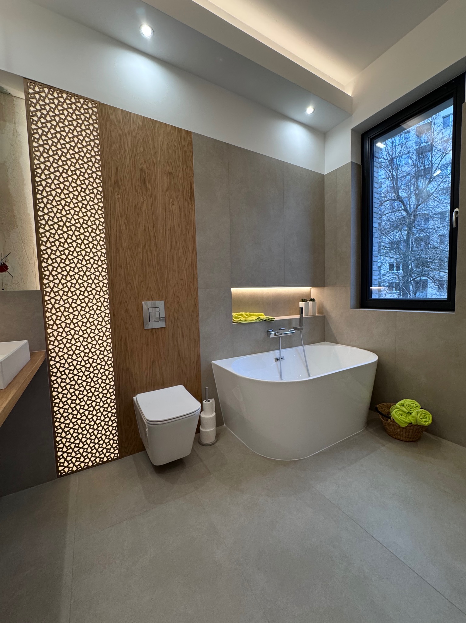 Nowoczesna łazienka z wolnostojącą wanną, drewnianą ścianką działową z ażurowym wzorem i dużym oknem z widokiem na drzewo. Minimalistyczny design.