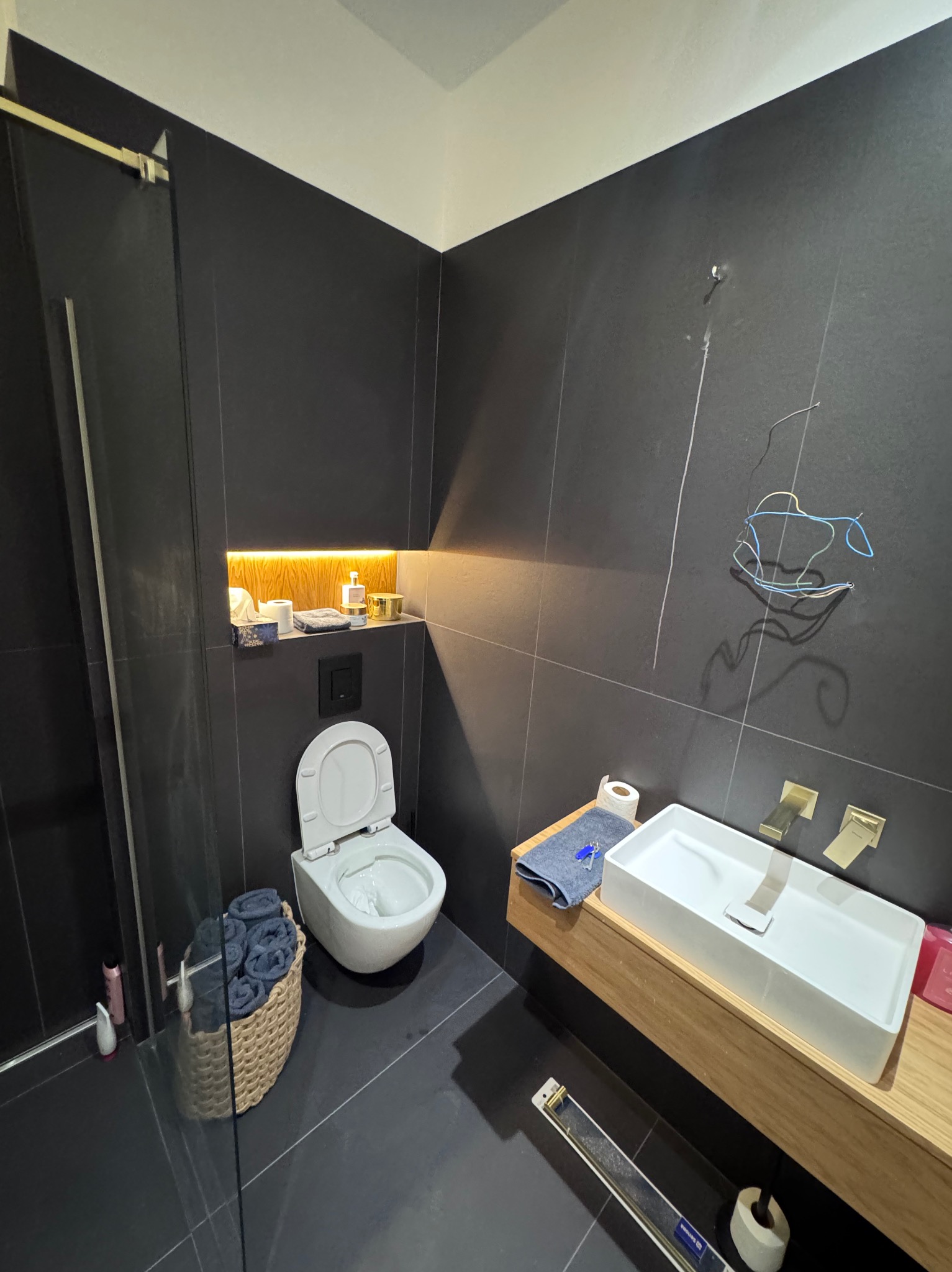 Ciemna łazienka z płytkami, podświetloną wnęką, umywalką na drewnianym blacie i otwartą toaletą. Widoczne kable instalacji elektrycznej.