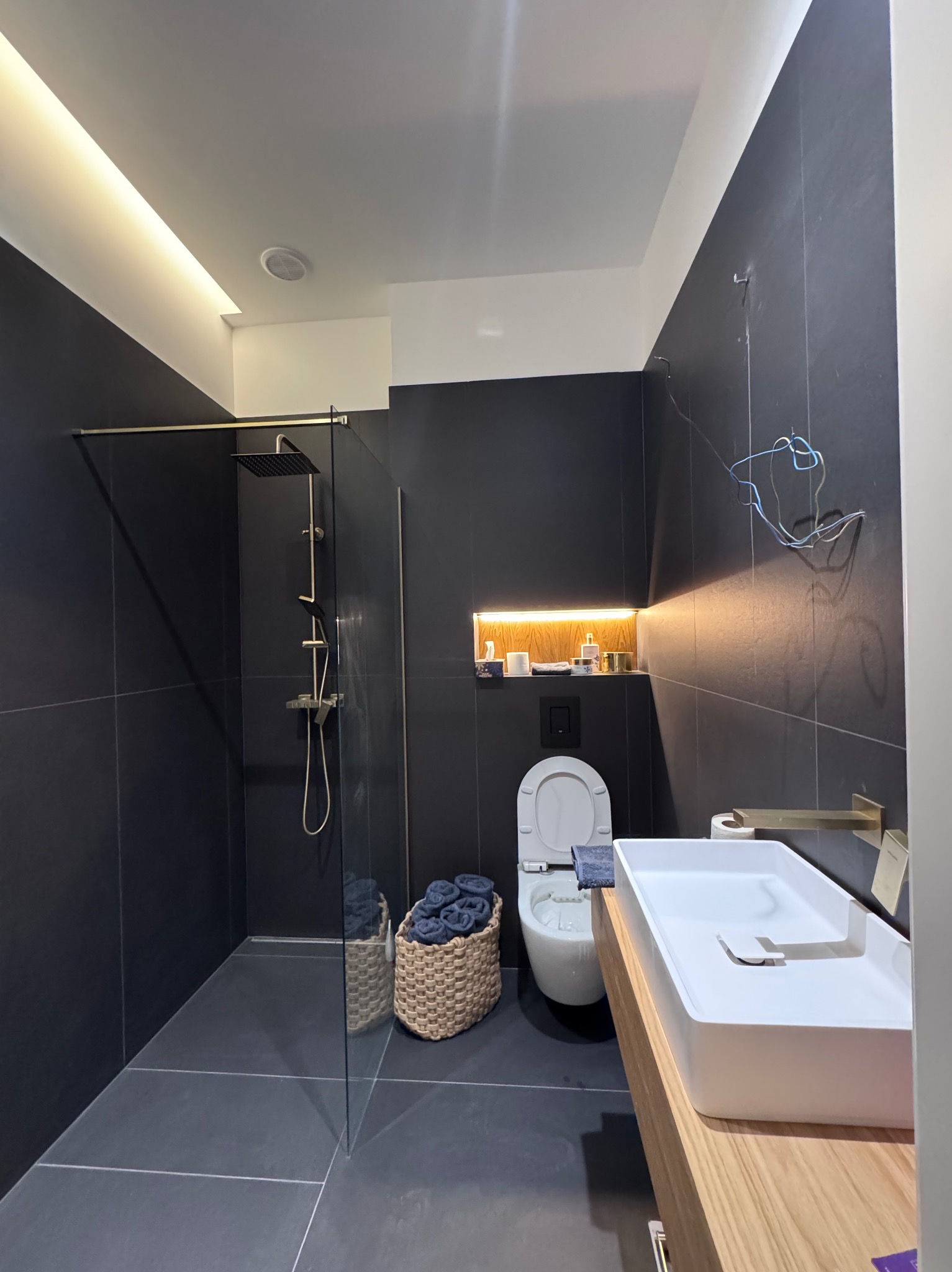 Ciemna łazienka z kabiną prysznicową typu walk-in, wiszącą toaletą i umywalką nablatową. Widoczne oświetlenie ledowe i kosz z ręcznikami.