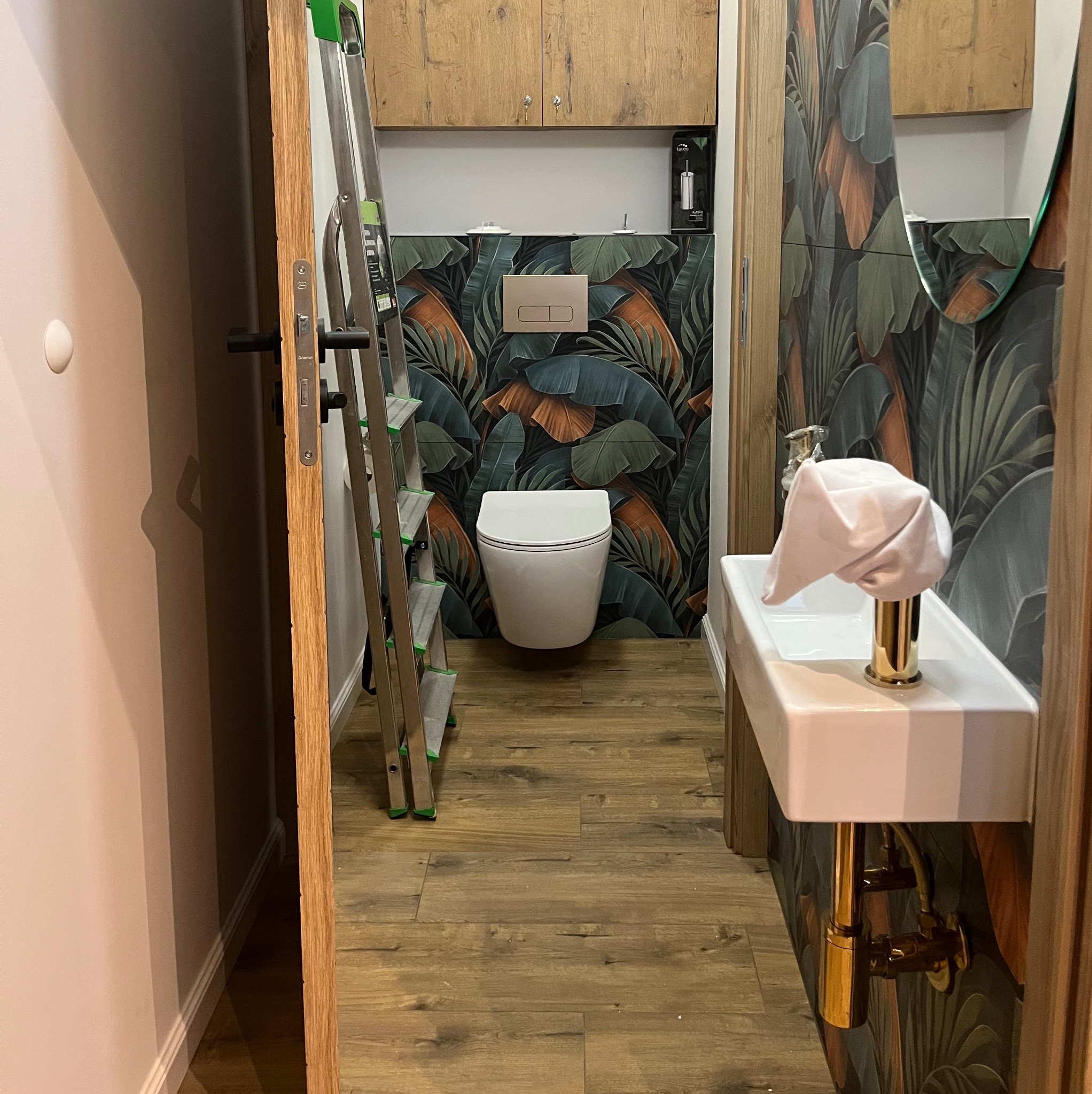 Wykończona łazienka z motywem tropikalnym: umywalka z baterią w kolorze złota, wisząca toaleta i drabina malarska w tle. Widok zza futryny.