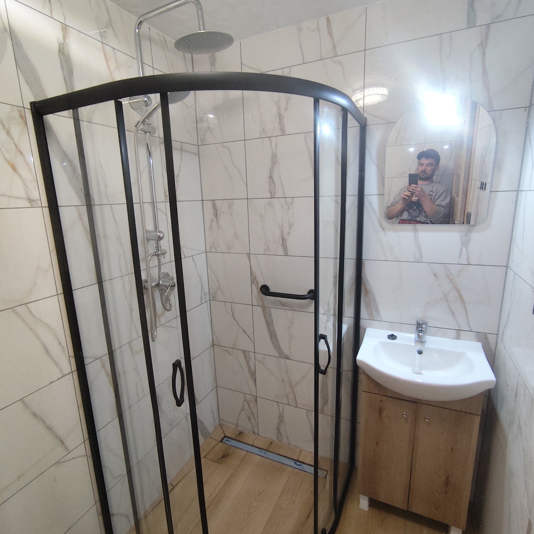Wykończona łazienka: kabina prysznicowa z czarną ramą, marmurowe płytki, umywalka z szafką. Widoczne odbicie osoby robiącej zdjęcie w lustrze.