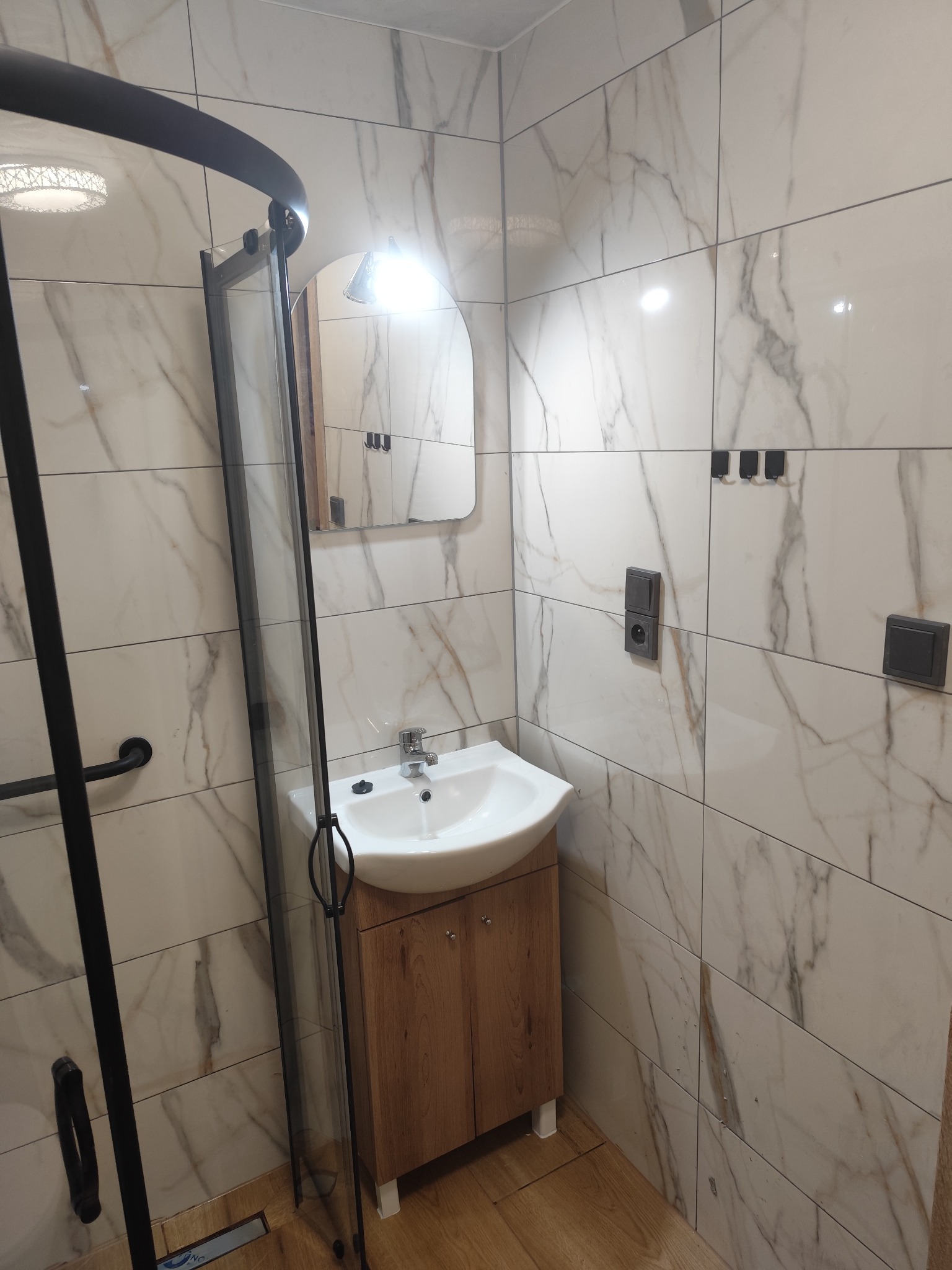 Wykończona łazienka z kabiną prysznicową typu walk-in, umywalką z szafką i lustrem. Marmurowe płytki na ścianach. Nowoczesny design. Widoczne czarne akcenty.