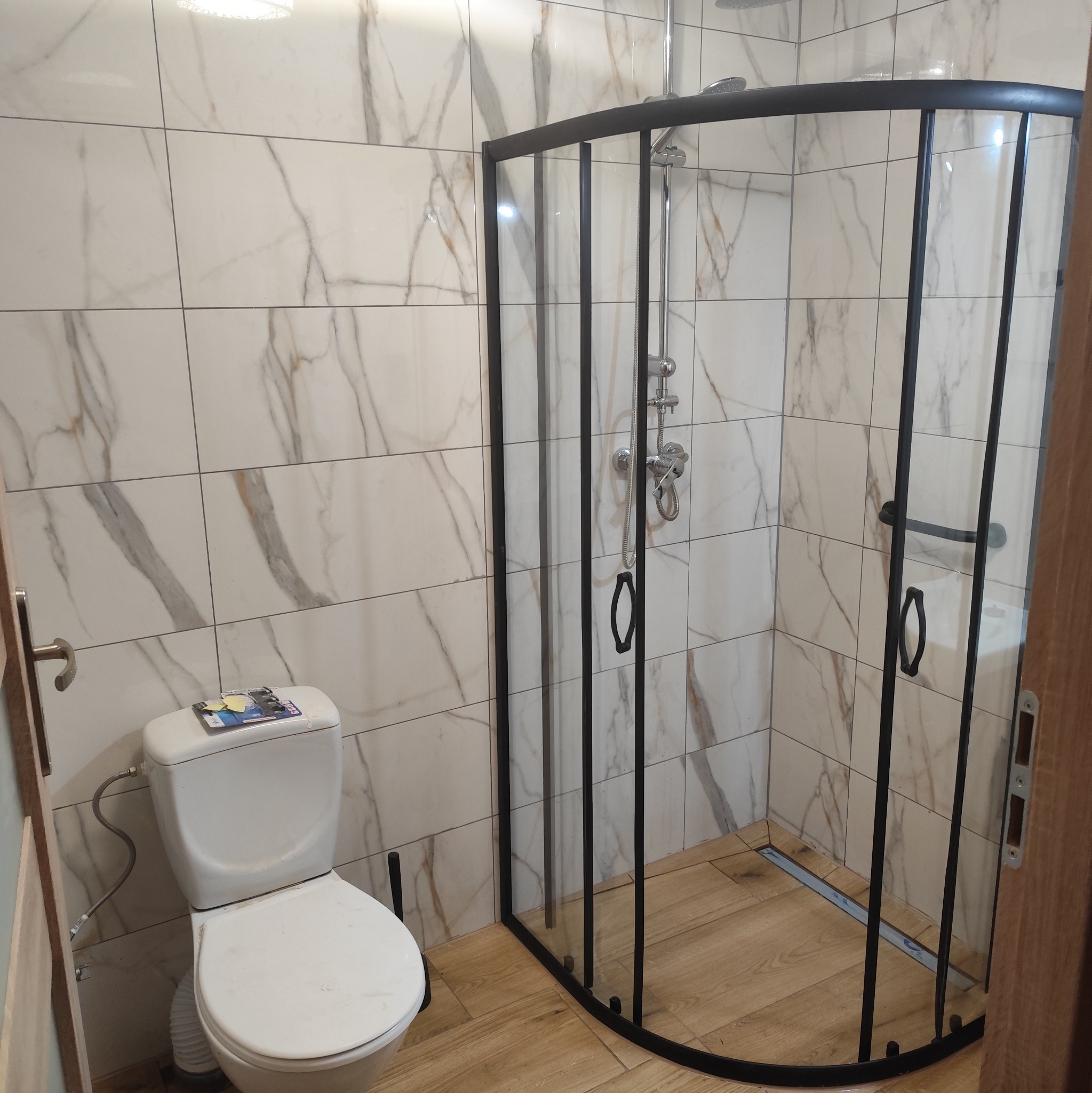 Nowoczesna kabina prysznicowa z czarnymi profilami w łazience wykończonej płytkami imitującymi marmur. Widoczna toaleta i drewnopodobna podłoga.