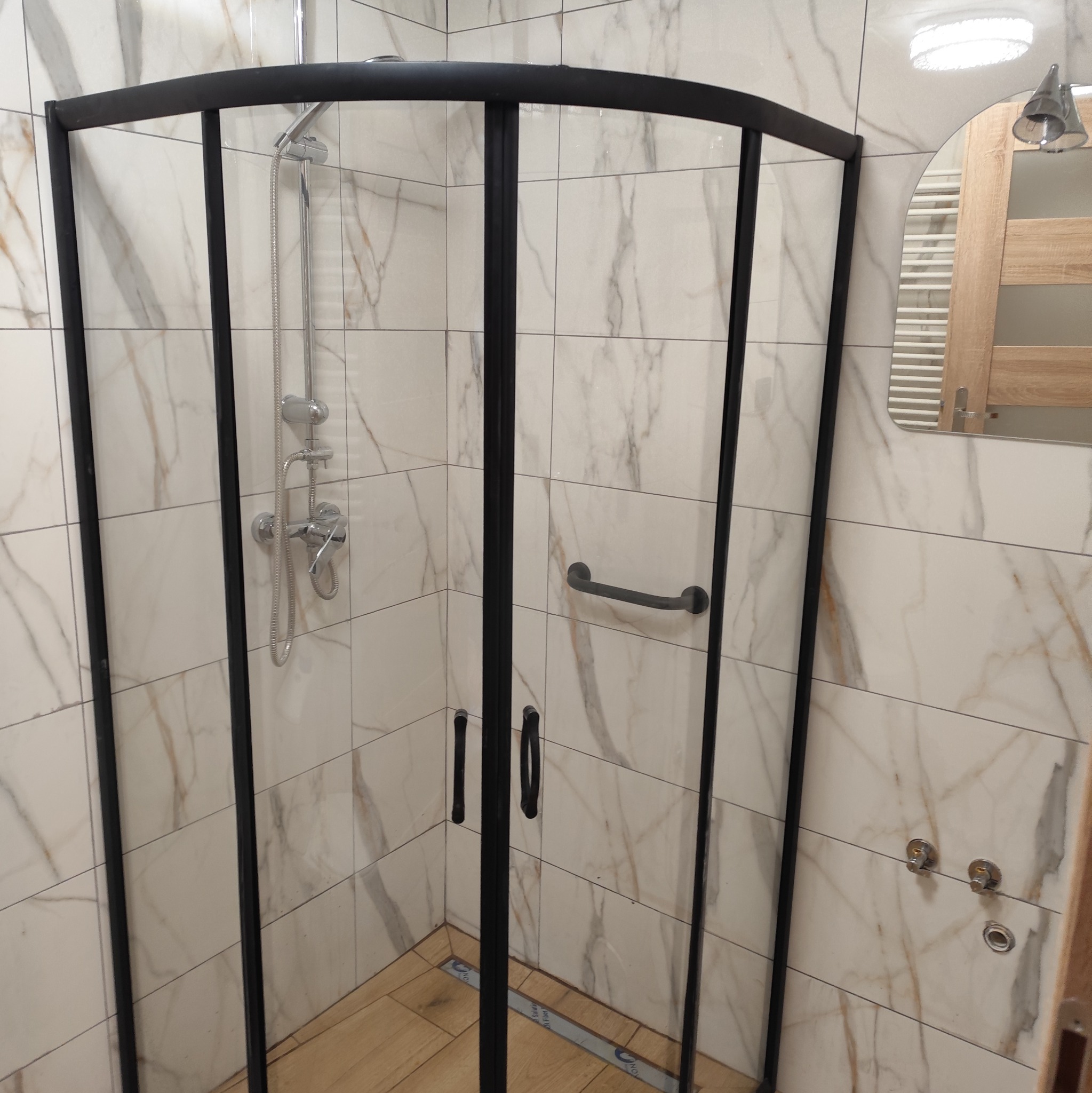 Kabina prysznicowa z czarnymi profilami na tle marmurowych płytek w łazience. Widoczny odpływ liniowy i armatura. Na ścianie lustro z grzejnikiem.