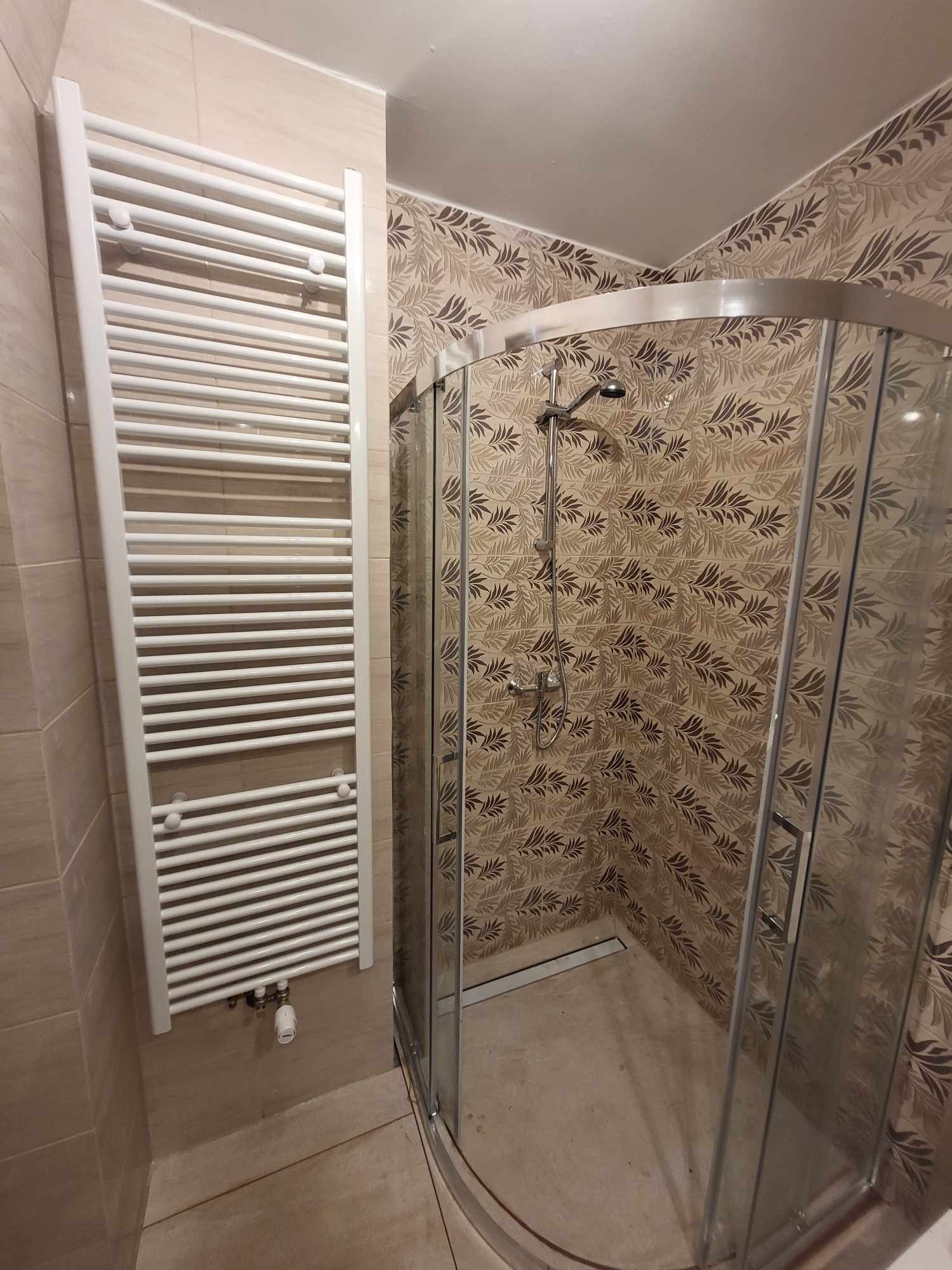 Wykończona łazienka z kabiną prysznicową i grzejnikiem drabinkowym. Beżowe płytki z motywem liści na ścianach. Nowoczesny design, widok na wnętrze kabiny.