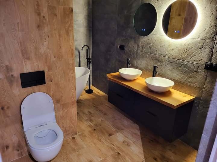 Nowoczesna łazienka z drewnianymi akcentami: płytki imitujące drewno, podwójna umywalka na blacie i okrągłe lustra z podświetleniem LED, minimalistyczny design.