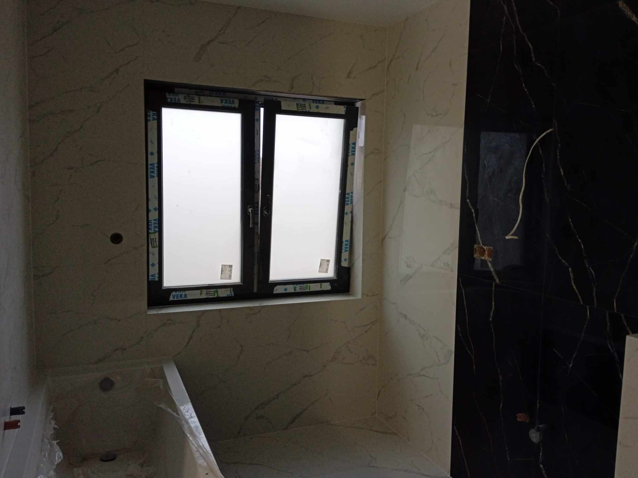 Wykończona łazienka z oknem i marmurowymi płytkami w odcieniach bieli i czerni. Widoczna wanna, instalacja elektryczna. Minimalistyczny design, nowoczesne wykończenie.