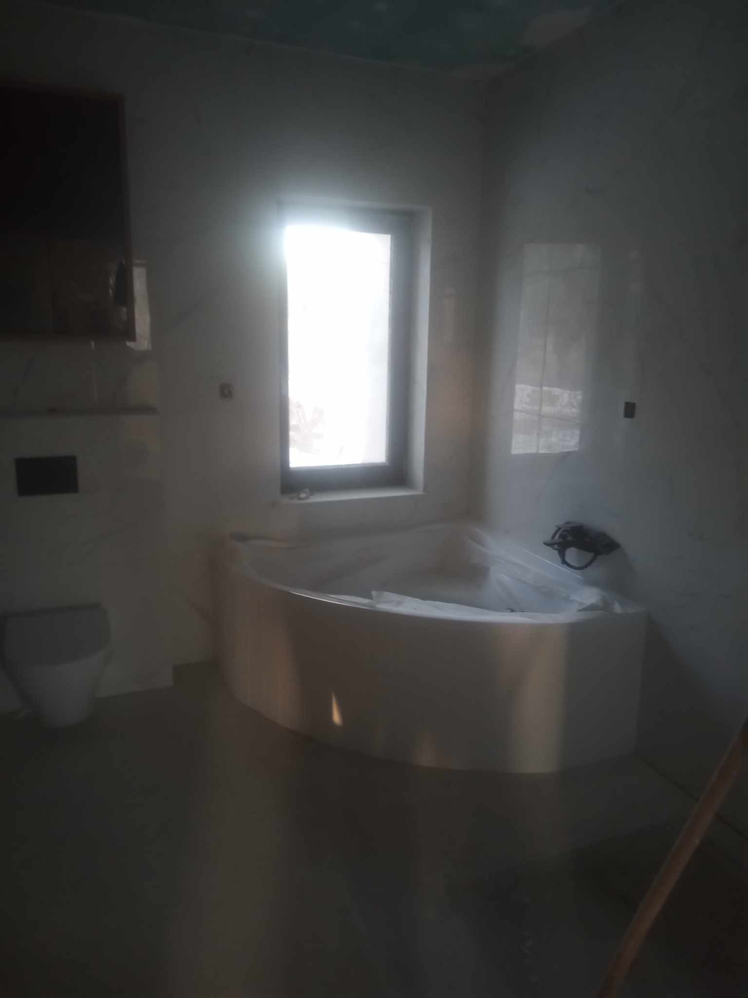 Jasna łazienka z wanną narożną przy oknie. Widoczne białe płytki na ścianach, częściowo zakryta wanna folią ochronną, umywalka i fragment drabiny w rogu.