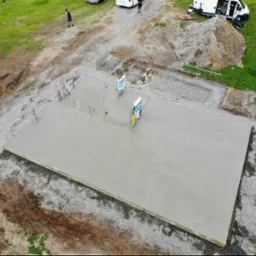 Świeżo wylany fundament pod budowę domu z wystającymi elementami instalacji, widok z góry. Na budowie obecni robotnicy i sprzęt.