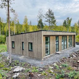 RR Scandic House - Nowoczesny, parterowy dom z drewnianą elewacją i dużymi oknami, otoczony zielenią i żwirowym podłożem. W tle widoczny las i palety budowlane.