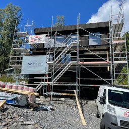 RR Scandic House - Czarny dom modułowy w trakcie budowy, otoczony rusztowaniami i materiałami budowlanymi. Na pierwszym planie samochód dostawczy z logo firmy budowlanej.