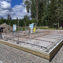 RR Scandic House - Zbrojona płyta fundamentowa z instalacjami pod dom, widok z boku na tle zieleni. Elementy szalunku z drewna. Na pierwszym planie żwir.