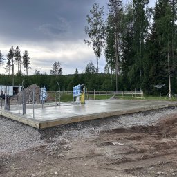 RR Scandic House - Wylany fundament pod budowę domu na działce z drzewami. Widoczne wystające rury, szalunek i gruz. W tle zaparkowany biały bus i ogrodzenie.