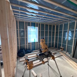 RR Scandic House - Wnętrze w trakcie budowy domu szkieletowego: widoczne drewniane ramy ścian, izolacja, okno i piła stołowa DeWalt na statywie. Etap wznoszenia konstrukcji.