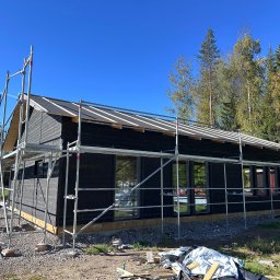 RR Scandic House - Czarny dom w trakcie budowy, otoczony rusztowaniem na tle lasu i błękitnego nieba. Widoczny fragment dachu i okna. Na pierwszym planie gruz i materiały budowlane.