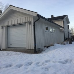 RR Scandic House - Biały dom szkieletowy z garażem w zimowym krajobrazie. Widoczne detale wykończenia, takie jak oświetlenie zewnętrzne i rynny. Śnieg pokrywa teren wokół budynku.