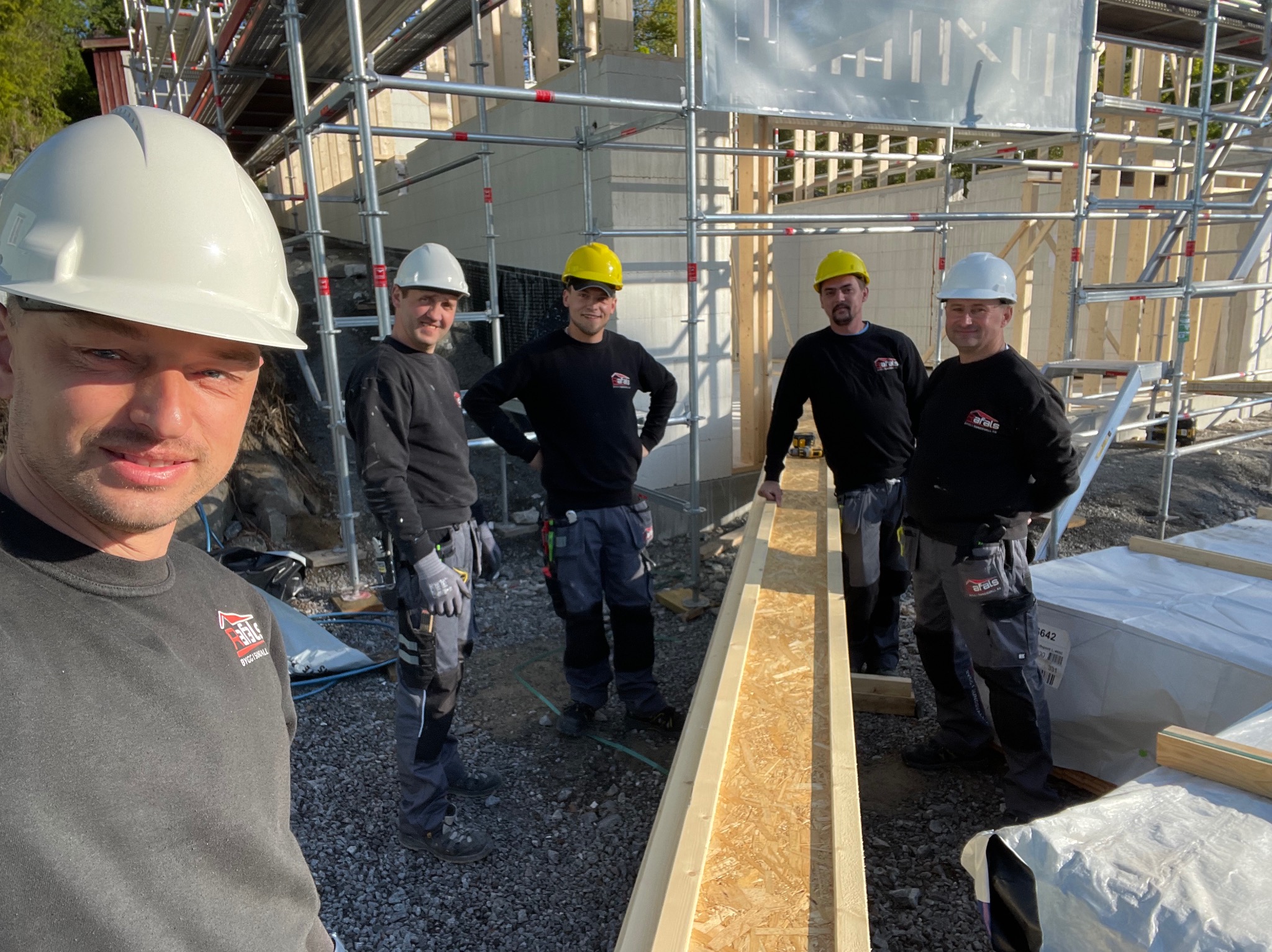 Ekipa budowlana w kaskach pozuje na tle konstrukcji domu szkieletowego. Widoczne rusztowania i elementy drewniane. Selfie z budowy w słoneczny dzień.