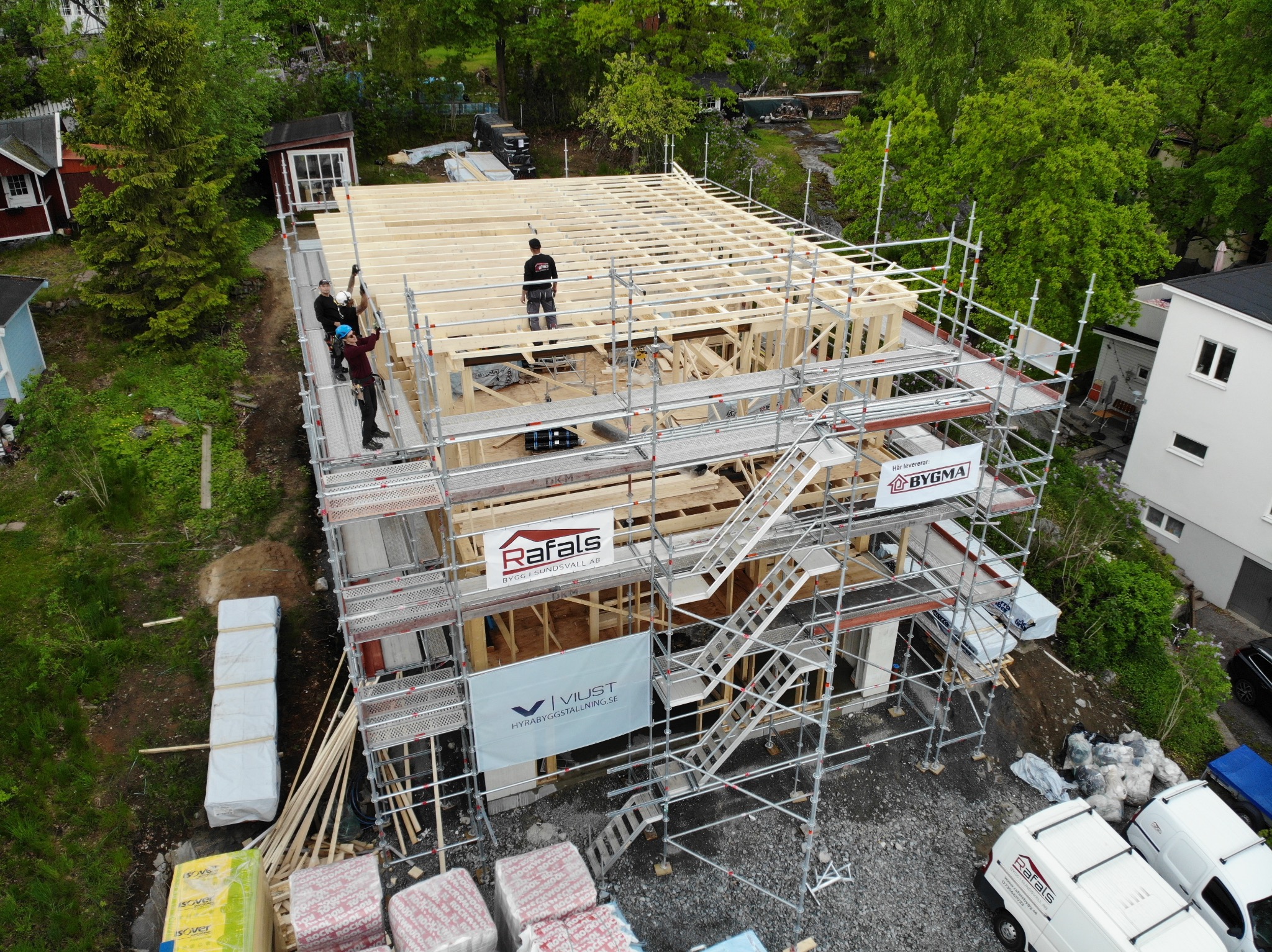Budowa domu w trakcie realizacji, widok z góry. Ekipa na rusztowaniu montuje drewnianą konstrukcję szkieletową. Na dole materiały budowlane i samochody dostawcze.