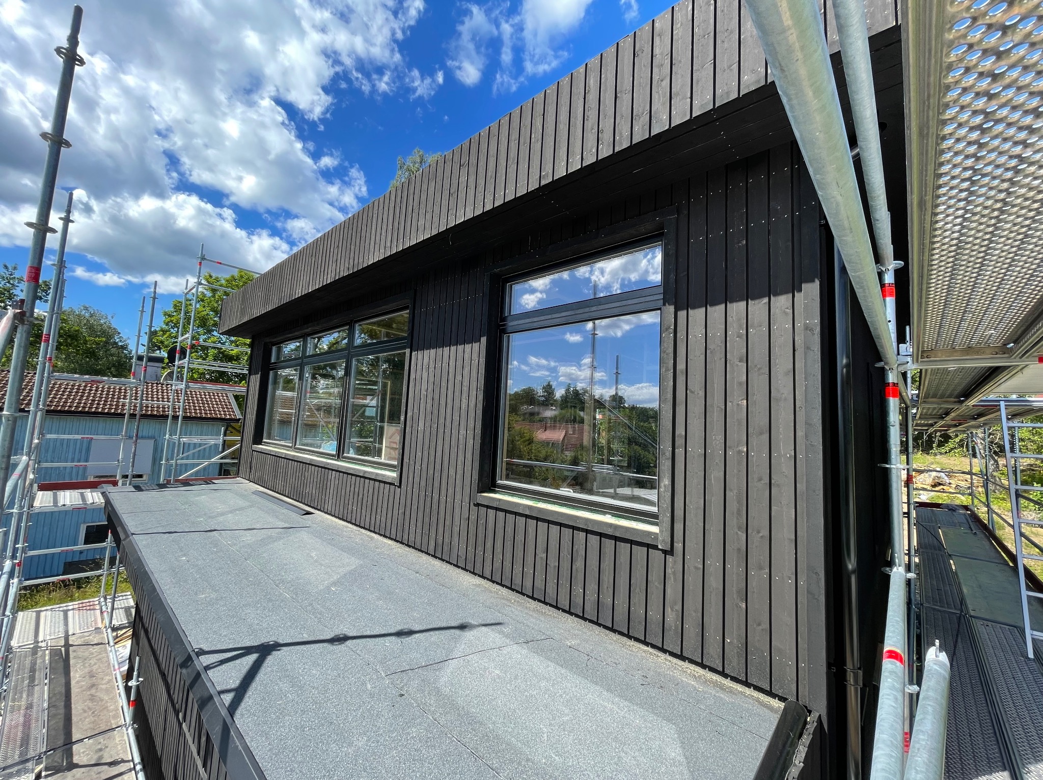 Ciemna, drewniana elewacja domu z widocznymi oknami i rusztowaniami. W oknach odbija się błękitne niebo z chmurami. Ujęcie z dołu, podkreślające nowoczesny design.