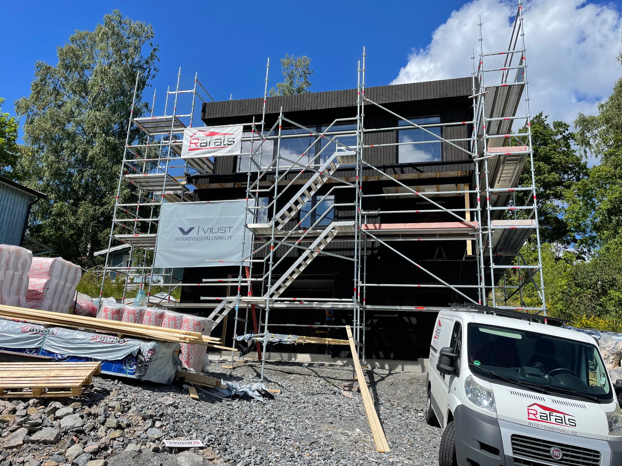 Czarny dom modułowy w trakcie budowy, otoczony rusztowaniami i materiałami budowlanymi. Na pierwszym planie samochód dostawczy z logo firmy budowlanej.