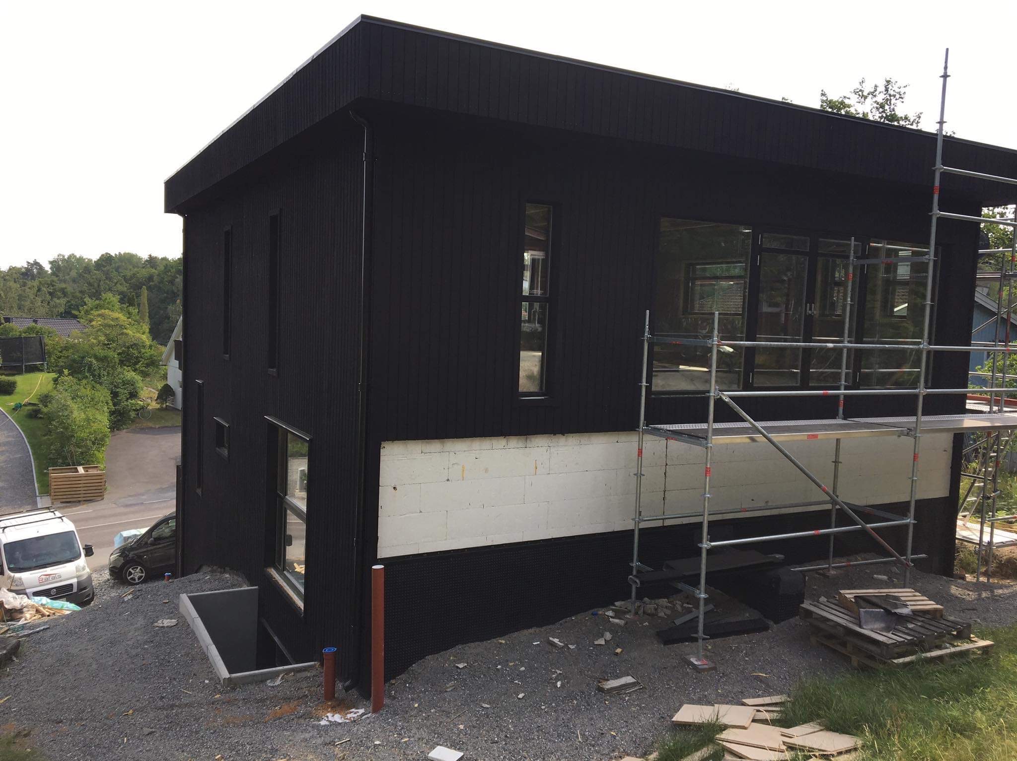 Czarny dom z drewnianą elewacją w trakcie budowy w Rątach. Widoczne rusztowania, okna i wejście do piwnicy. Część elewacji ocieplona styropianem.
