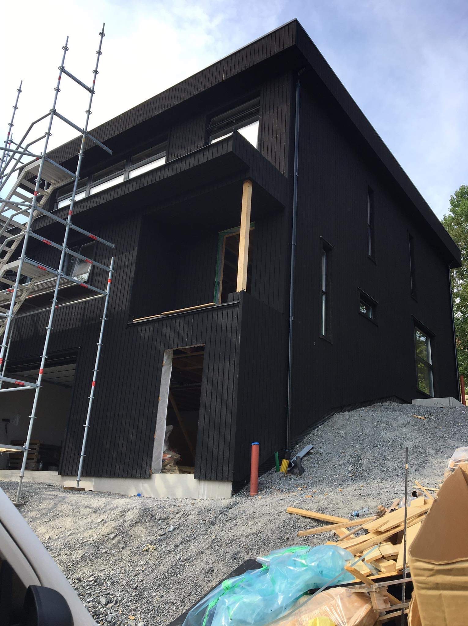 Czarny, dwukondygnacyjny dom z drewnianą elewacją w trakcie budowy. Widoczne rusztowania, gruz i materiały budowlane. Ujęcie z dołu, podkreślające nowoczesną bryłę.