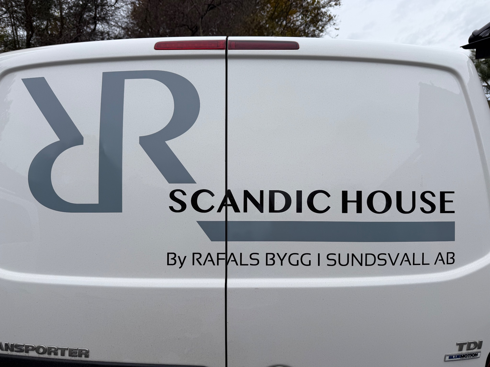 Tył białego samochodu dostawczego z szarym logo i napisem 'SCANDIC HOUSE By RAFALS BYGG I SUNDSVALL AB'. Widoczne oznaczenia modelu 'TRANSPORTER' i 'TDI'.