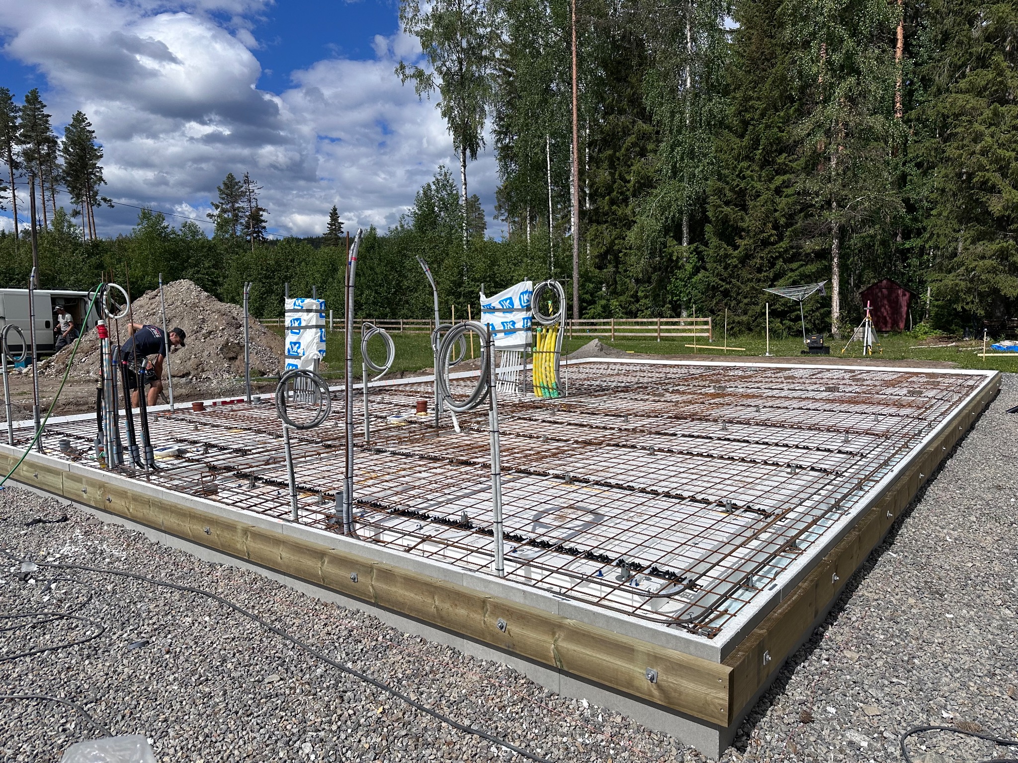 Zbrojona płyta fundamentowa z instalacjami pod dom, widok z boku na tle zieleni. Elementy szalunku z drewna. Na pierwszym planie żwir.