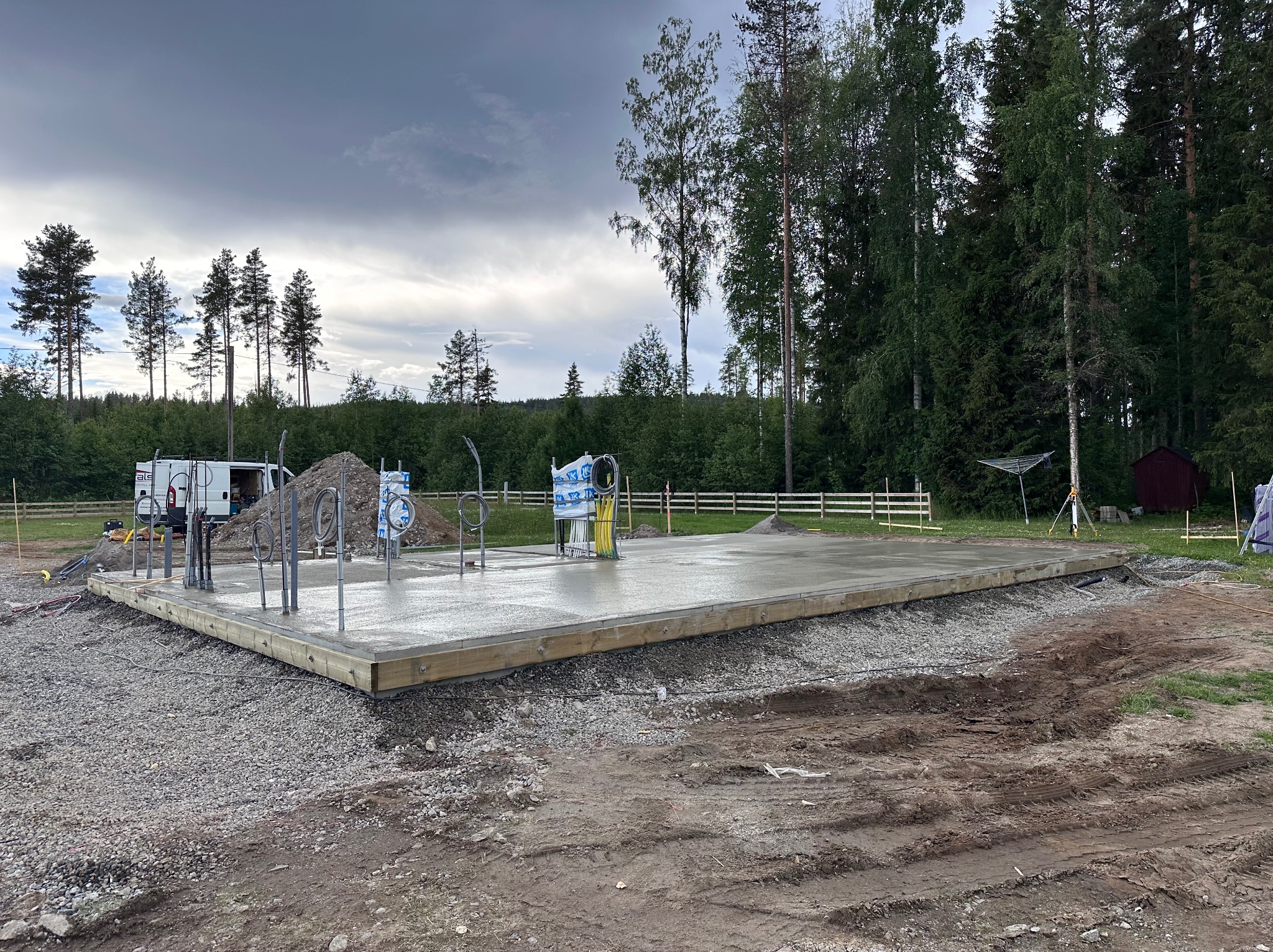 Wylany fundament pod budowę domu na działce z drzewami. Widoczne wystające rury, szalunek i gruz. W tle zaparkowany biały bus i ogrodzenie.