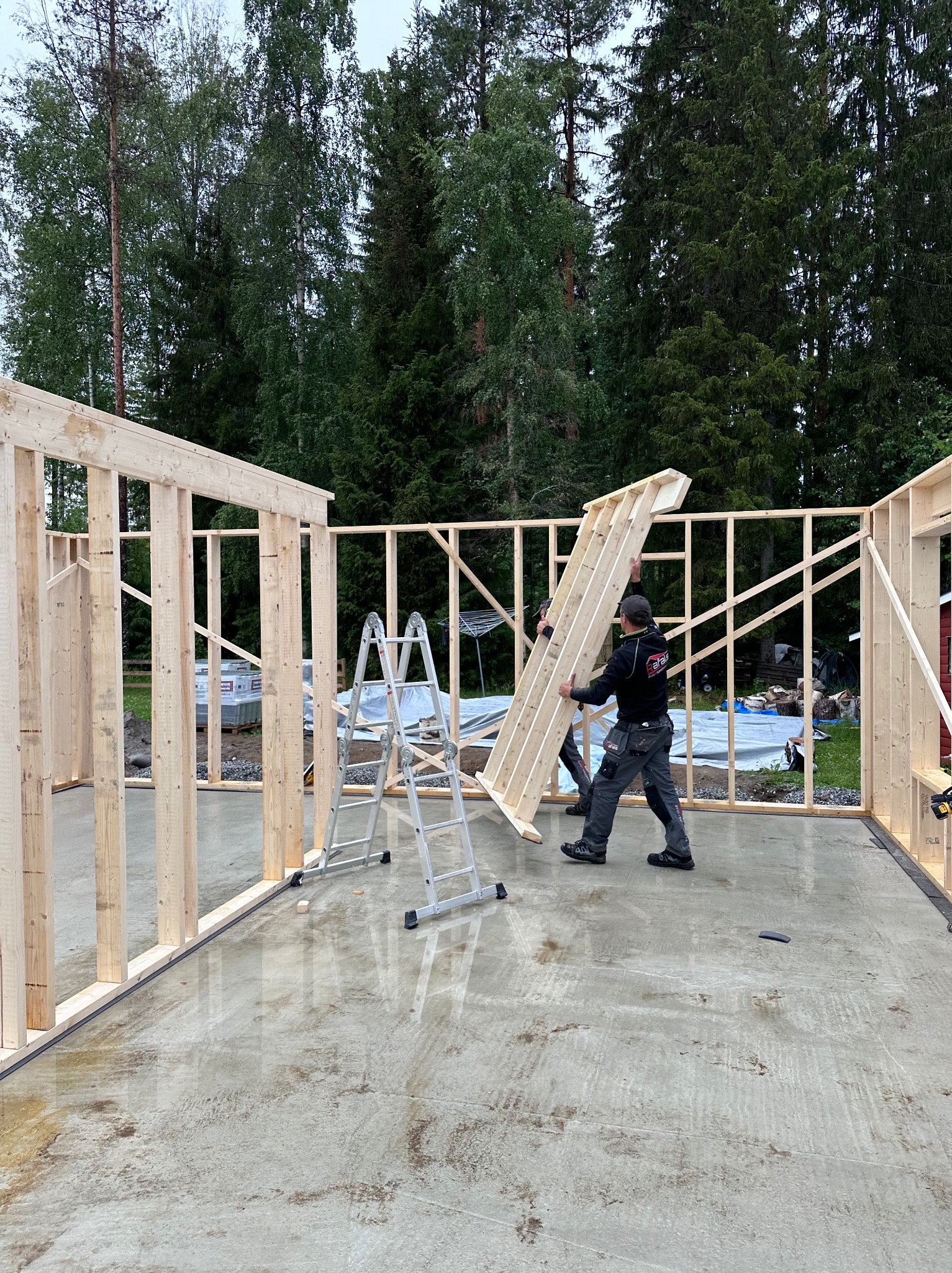 Montaż ściany szkieletowej z drewna na mokrym fundamencie. Dwóch pracowników w trakcie pracy. W tle las i drabina.