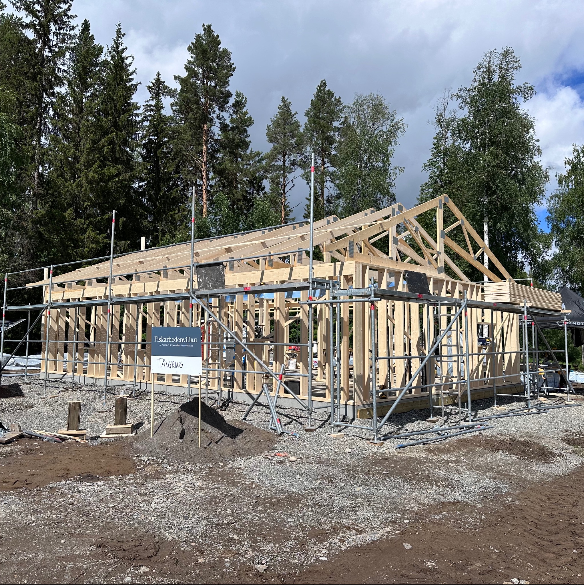 Budowa domu szkieletowego w lesie, widoczne rusztowania i konstrukcja drewniana. Tablica informacyjna z nazwą firmy budowlanej na pierwszym planie.