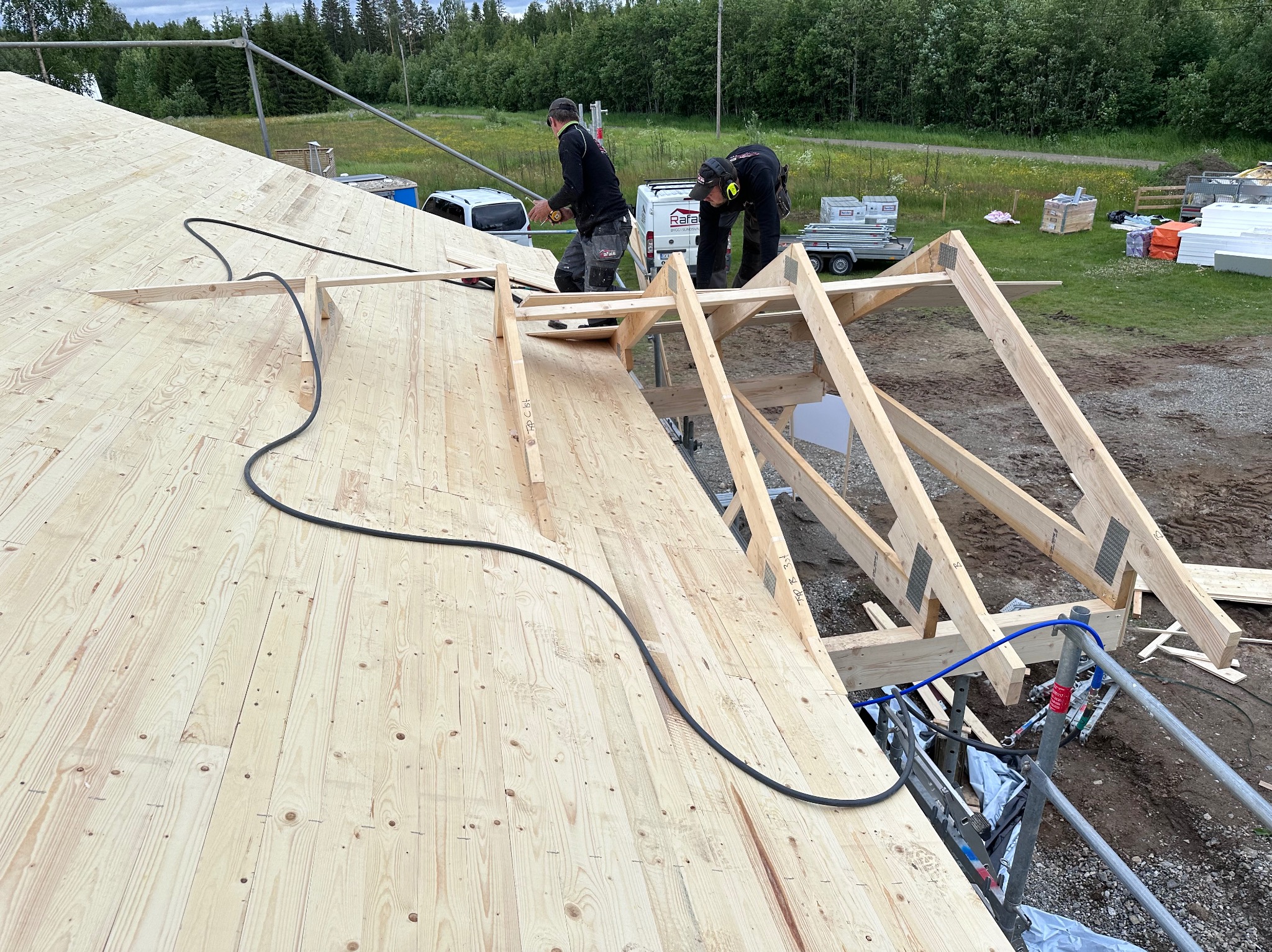 Montaż drewnianej konstrukcji dachu z widocznymi elementami więźby i deskowaniem. Dwóch pracowników w trakcie pracy, narzędzia i materiały budowlane w tle.