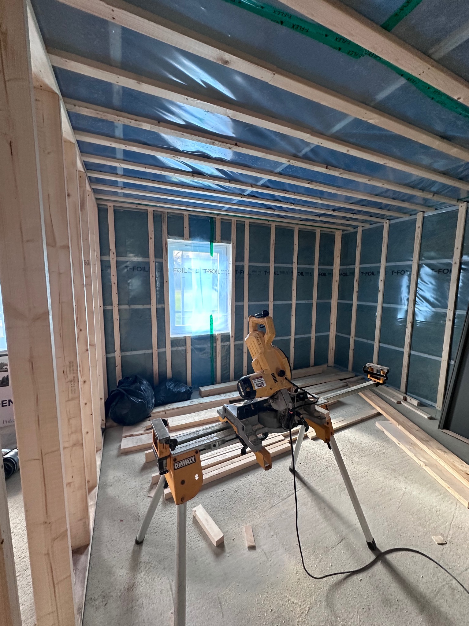 Wnętrze w trakcie budowy domu szkieletowego: widoczne drewniane ramy ścian, izolacja, okno i piła stołowa DeWalt na statywie. Etap wznoszenia konstrukcji.
