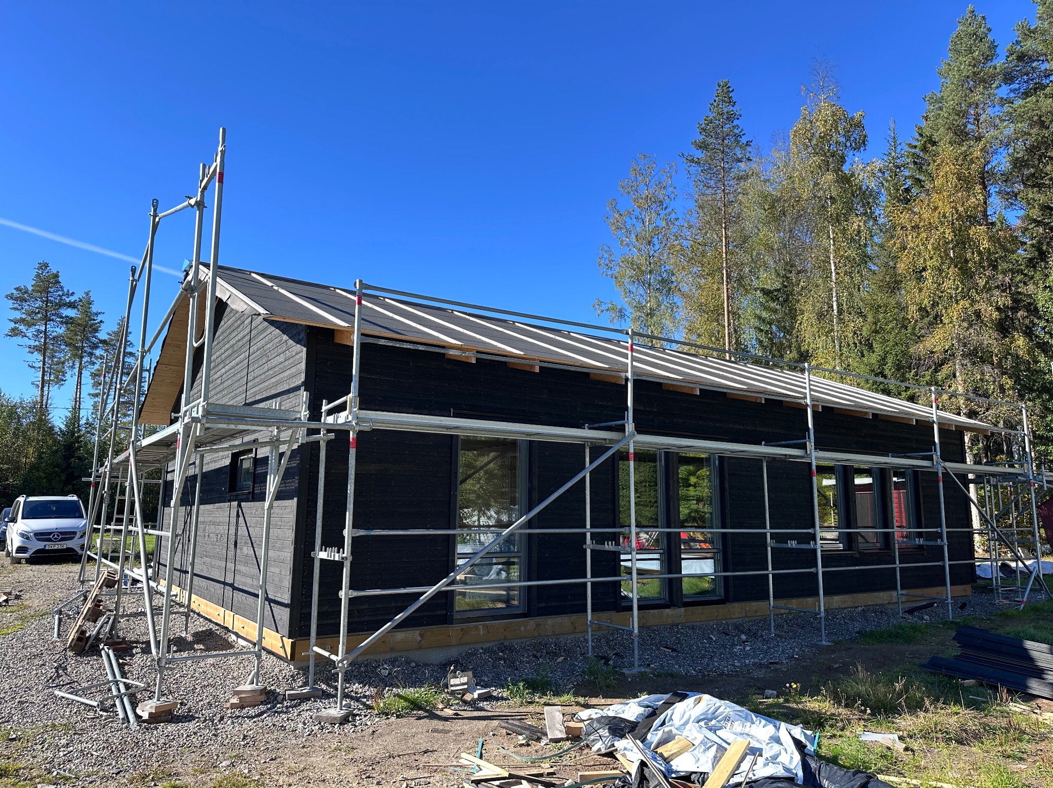 Czarny dom w trakcie budowy, otoczony rusztowaniem na tle lasu i błękitnego nieba. Widoczny fragment dachu i okna. Na pierwszym planie gruz i materiały budowlane.