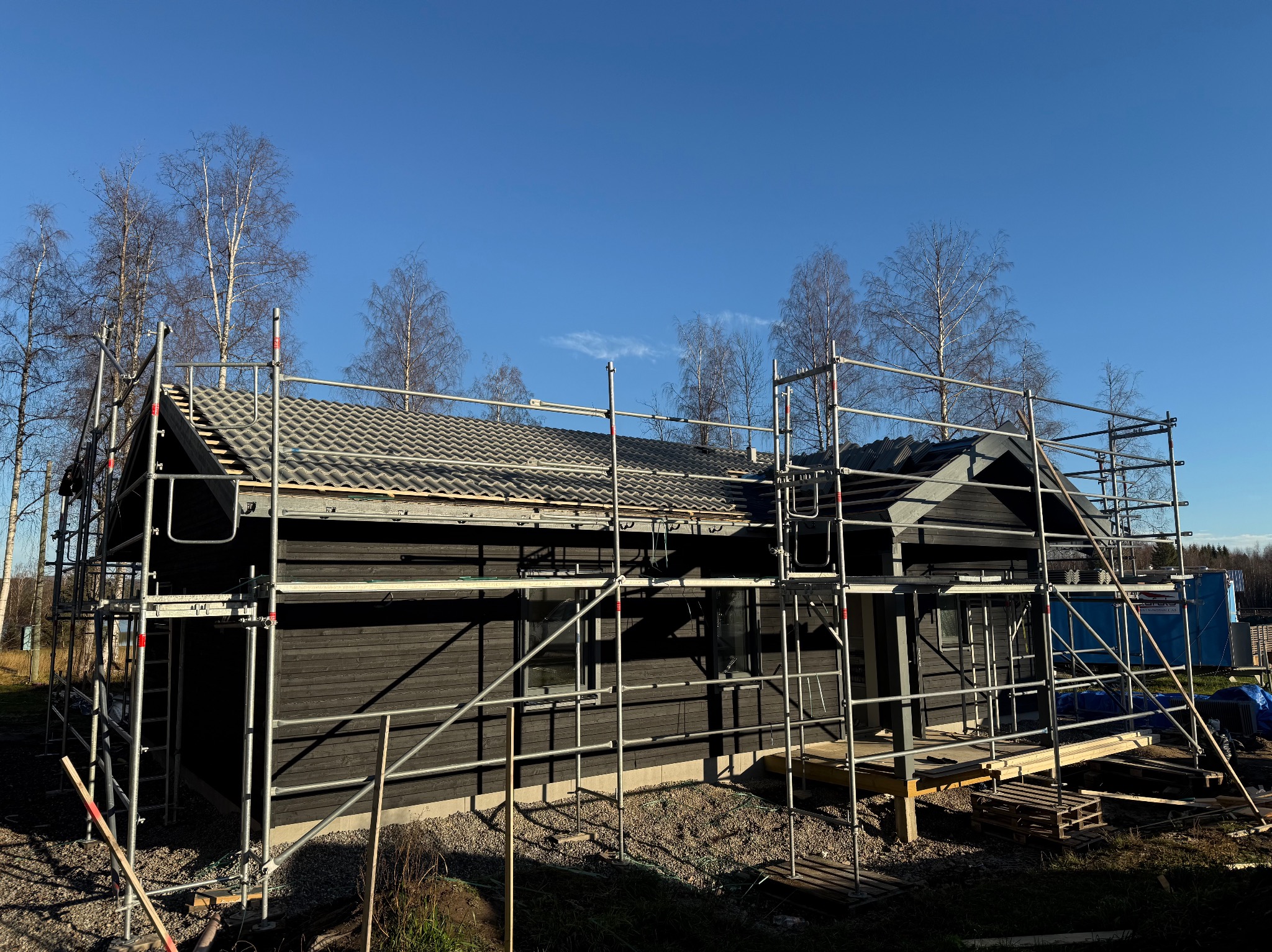 Ciemny dom szkieletowy w trakcie budowy, otoczony rusztowaniem. Widoczne szare dachówki i błękitne niebo w tle. Elementy konstrukcyjne i palety budowlane na pierwszym planie.