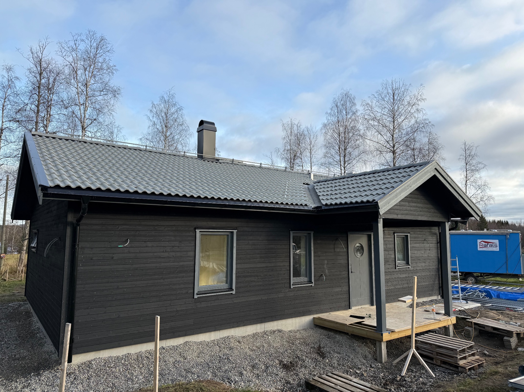 Czarny, parterowy dom z szarą dachówką i kominem. Widoczny fragment tarasu i niebieska przyczepa w tle. Ujęcie z poziomu gruntu, pochmurne niebo.
