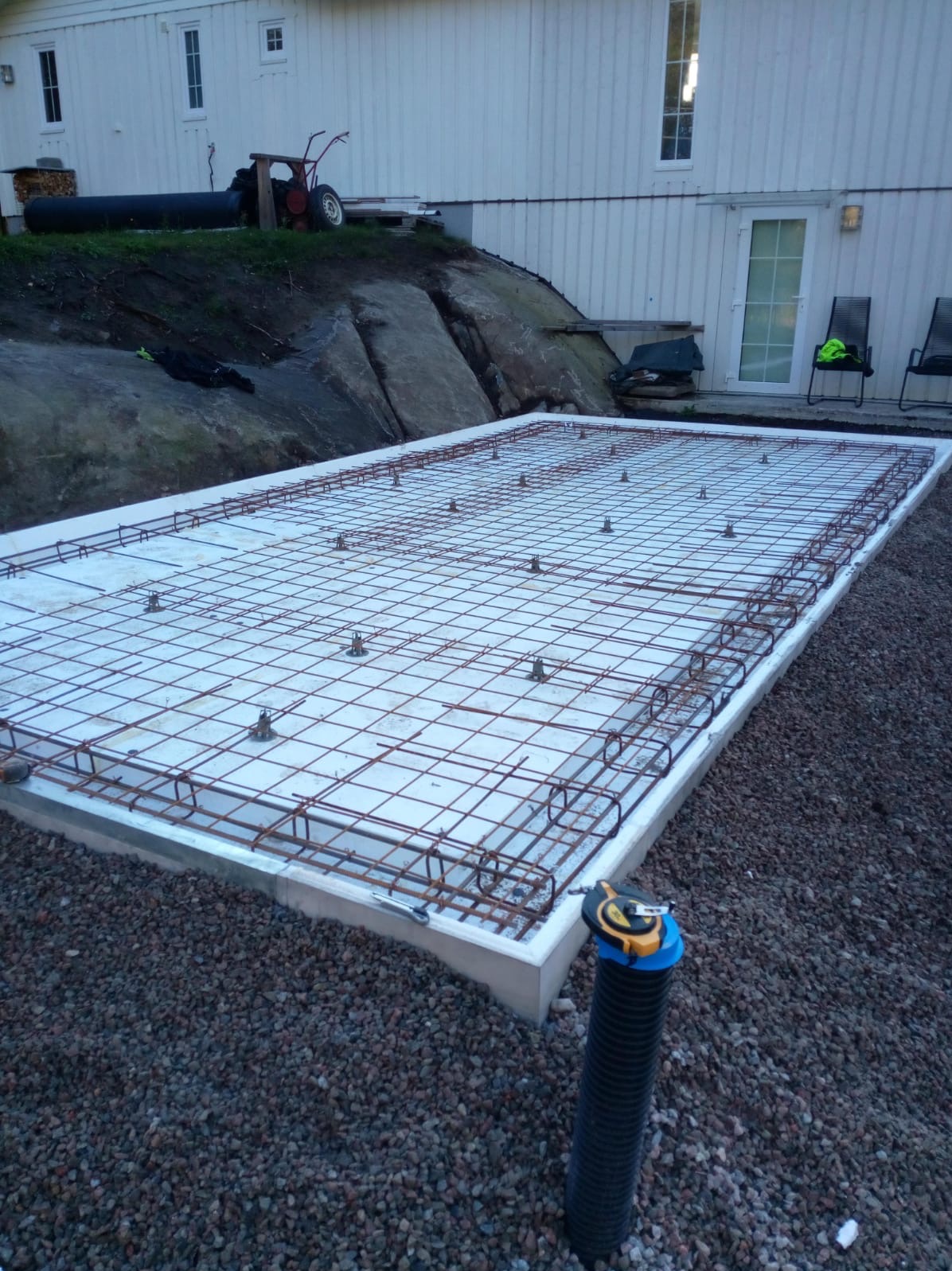 Zbrojona płyta fundamentowa z izolacją, przygotowana pod budowę domu. Widoczna siatka zbrojeniowa i elementy dystansowe. W tle budynek z białego sidingu.