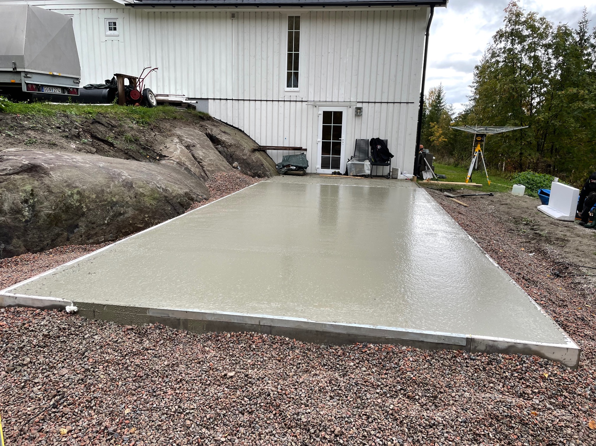 Świeżo wylana, gładka betonowa płyta fundamentowa z metalowym obramowaniem, przygotowana pod budowę domu na działce z widoczną skałą i białym budynkiem w tle.