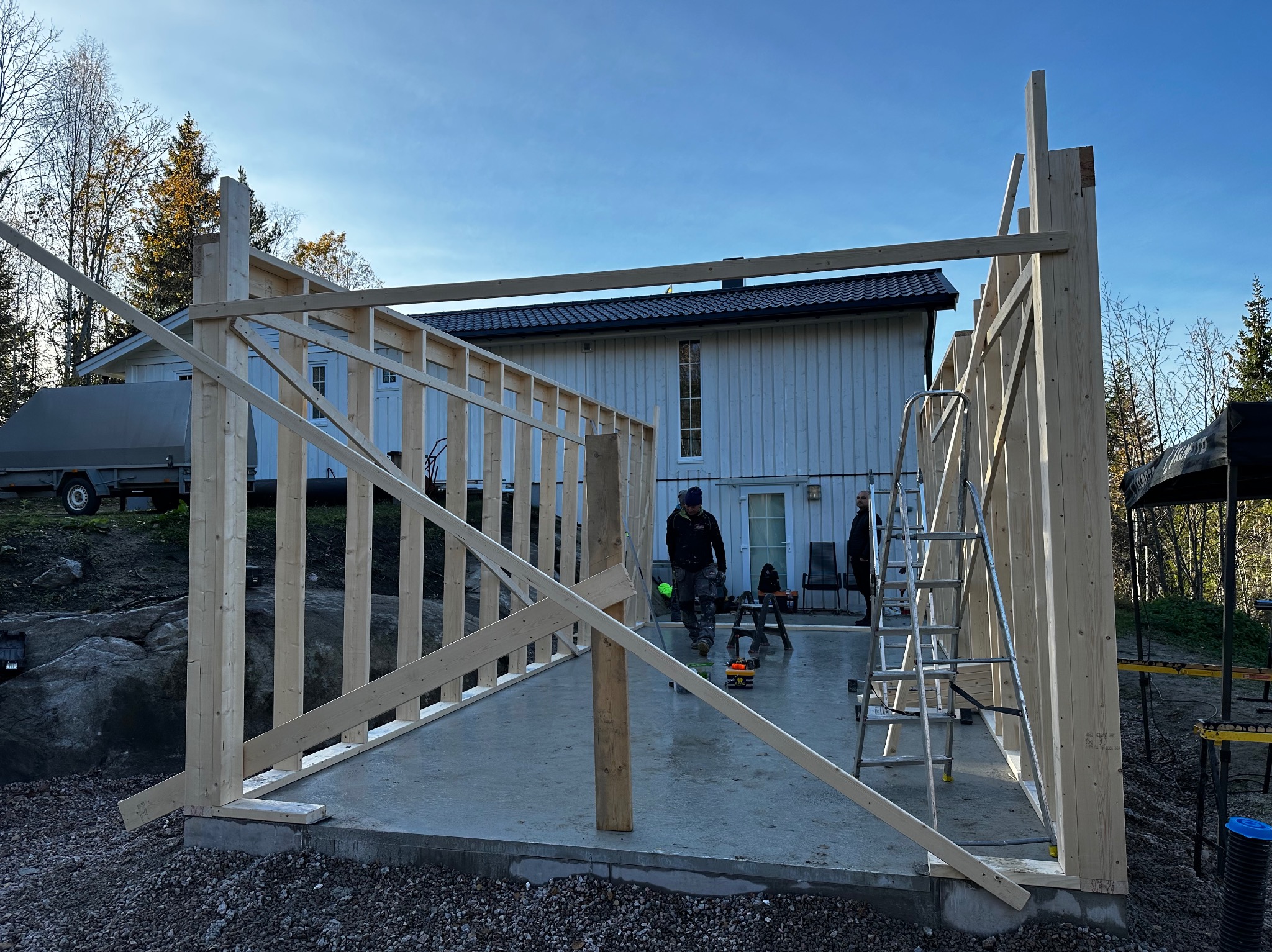 Wznoszenie konstrukcji szkieletowej budynku na fundamencie. Widoczni pracownicy i drabina. W tle dom z białymi ścianami i ciemnym dachem.