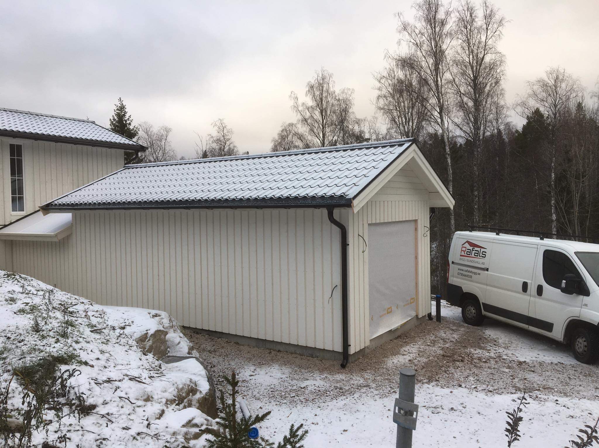 Biały, parterowy budynek z garażem w zimowej scenerii. Śnieg na dachu i ziemi. Obok zaparkowany biały bus z logo firmy budowlanej. Widoczne drzewa w tle.