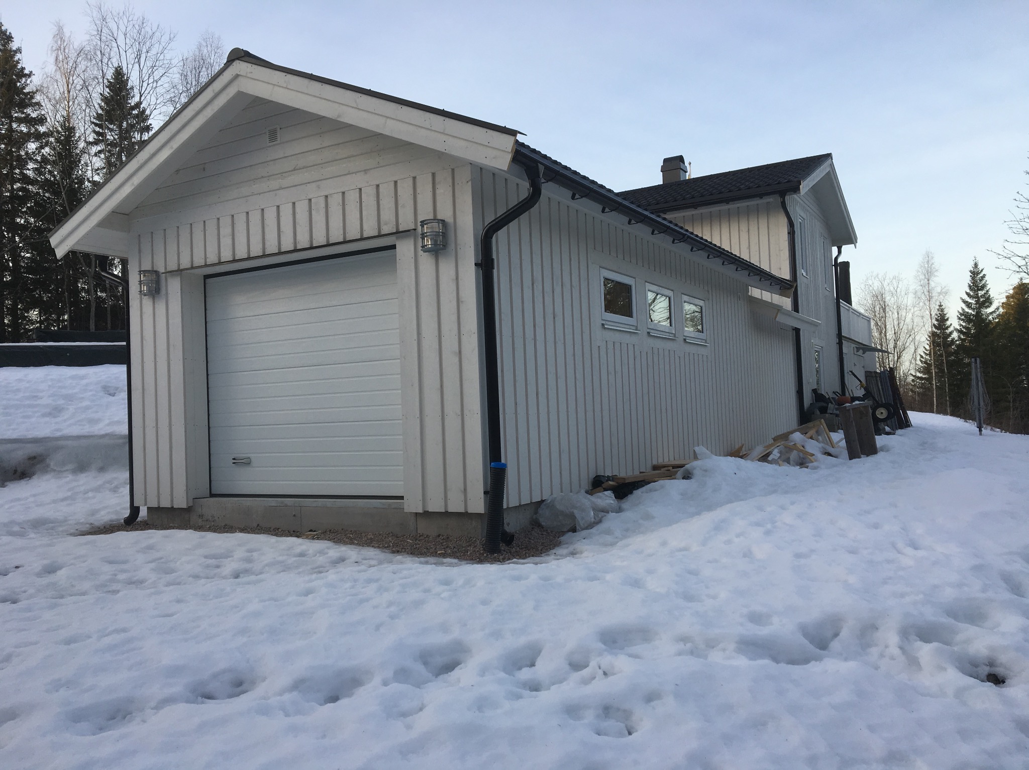 Biały dom szkieletowy z garażem w zimowym krajobrazie. Widoczne detale wykończenia, takie jak oświetlenie zewnętrzne i rynny. Śnieg pokrywa teren wokół budynku.