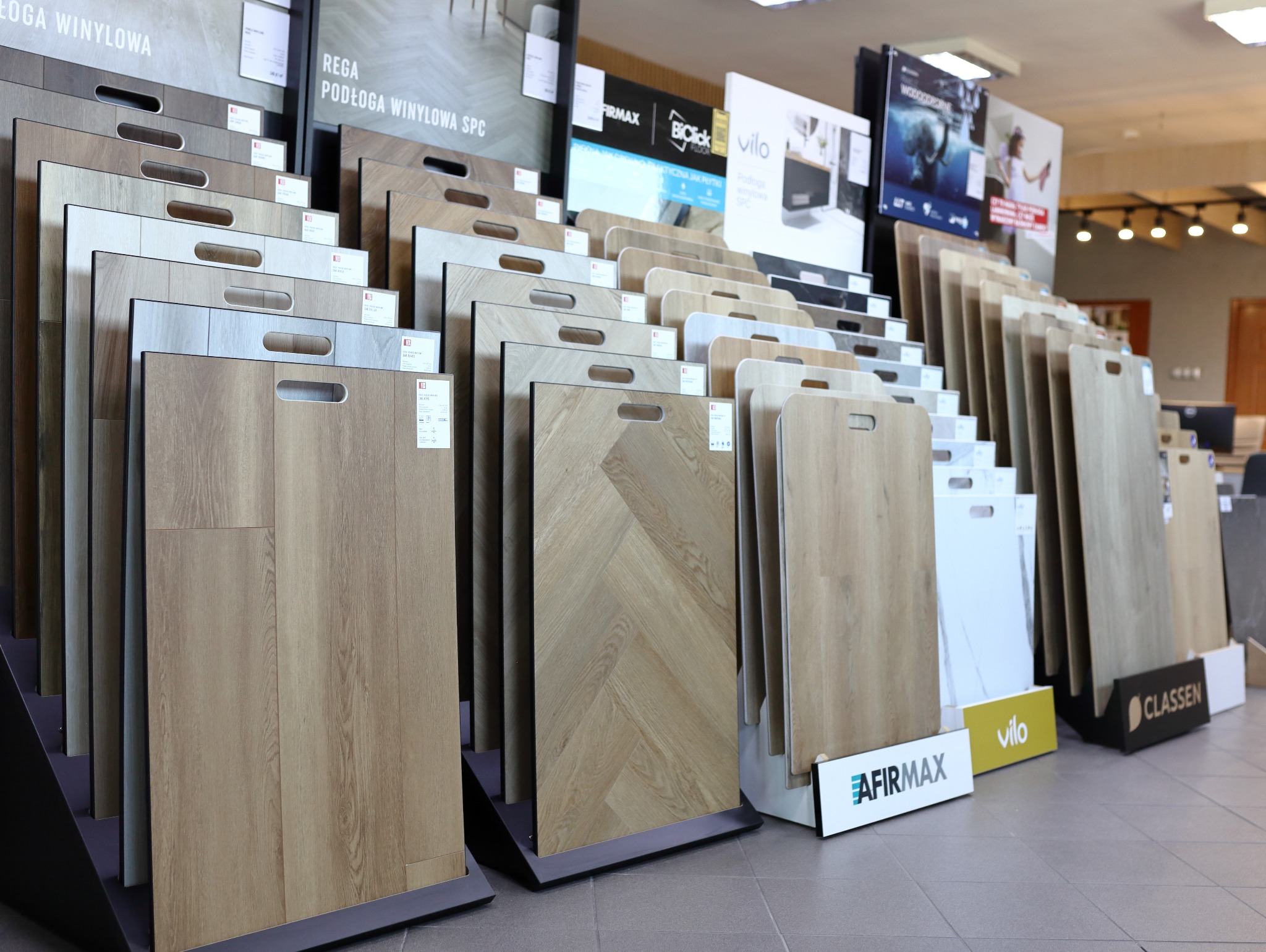 Ekspozycja paneli podłogowych w sklepie: różnorodne wzory, od klasycznych desek po jodełkę, z uchwytami do przenoszenia i logo producentów.