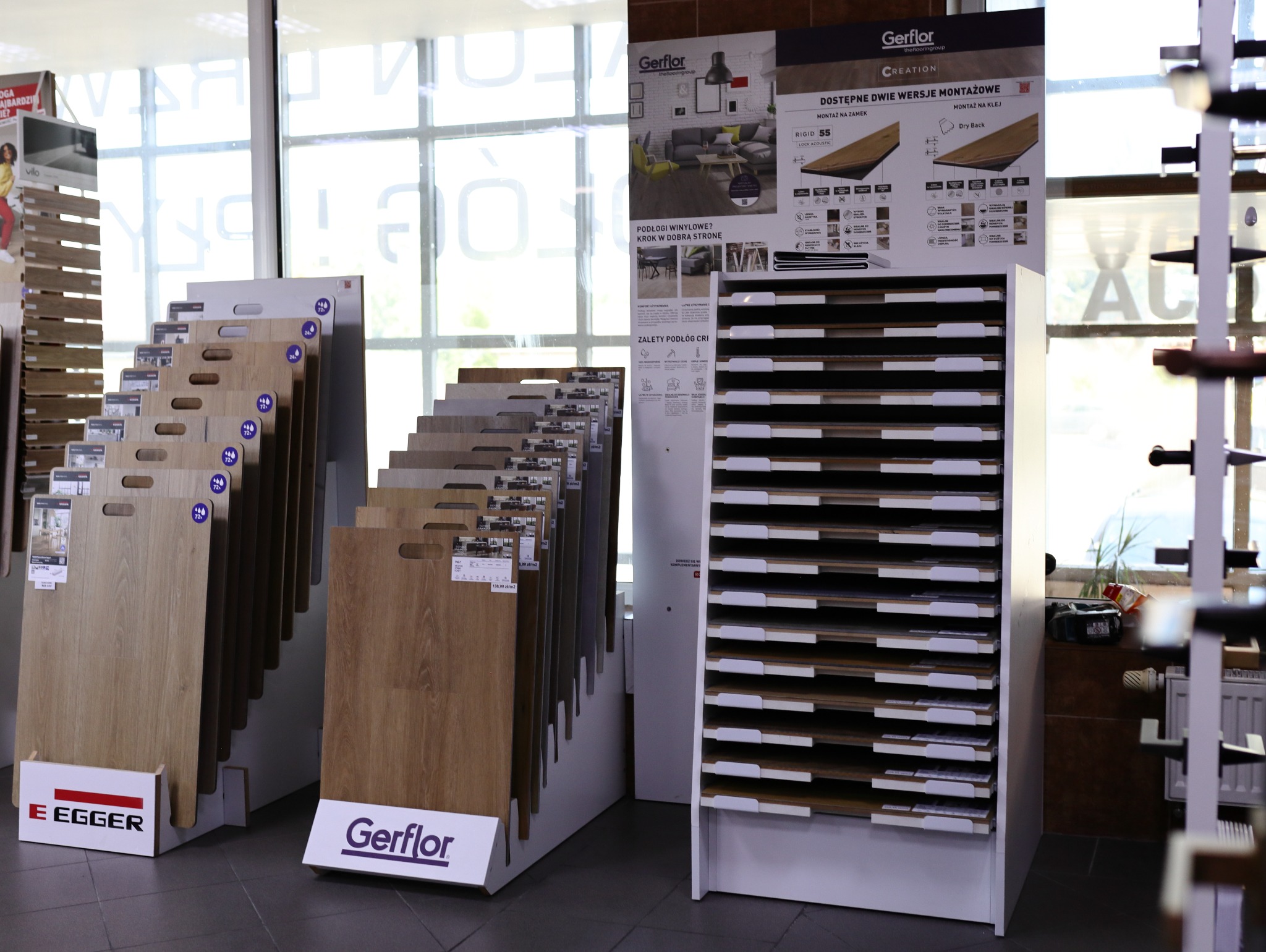 Ekspozycja paneli podłogowych marek Egger i Gerflor w sklepie. Widoczne wzorniki i reklamy, w tle okno z napisem. Prezentacja różnych odcieni i faktur paneli.