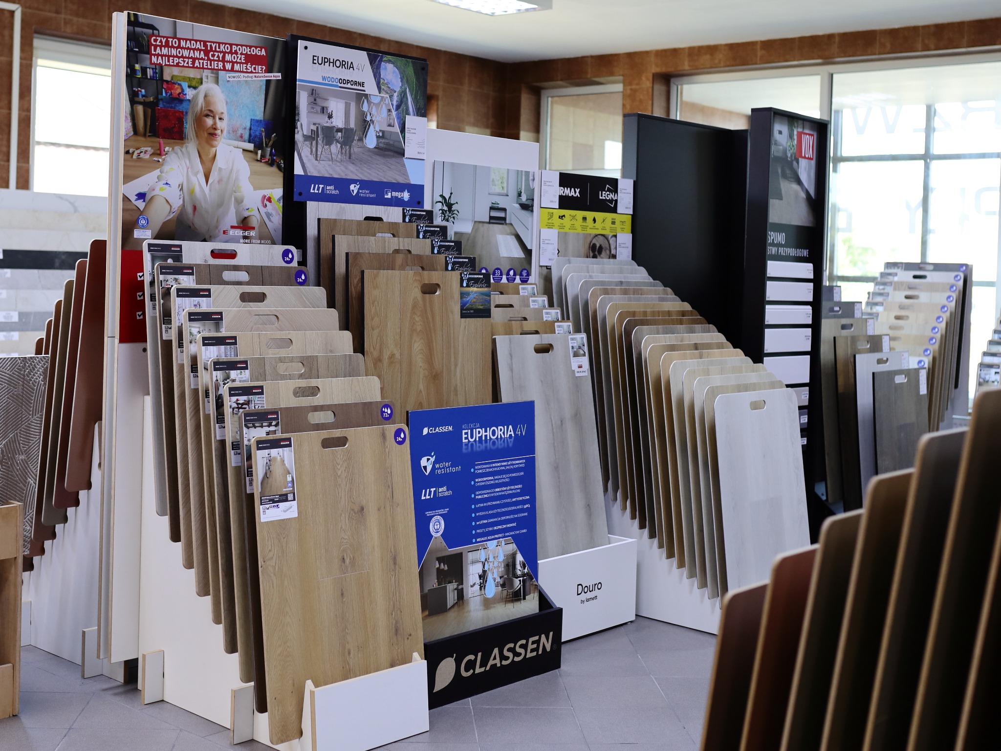 Ekspozycja paneli podłogowych w sklepie: różnorodne wzory i kolory, od jasnych po ciemne, z widocznymi oznaczeniami producentów i kolekcji, takich jak Classen Euphoria 4V.