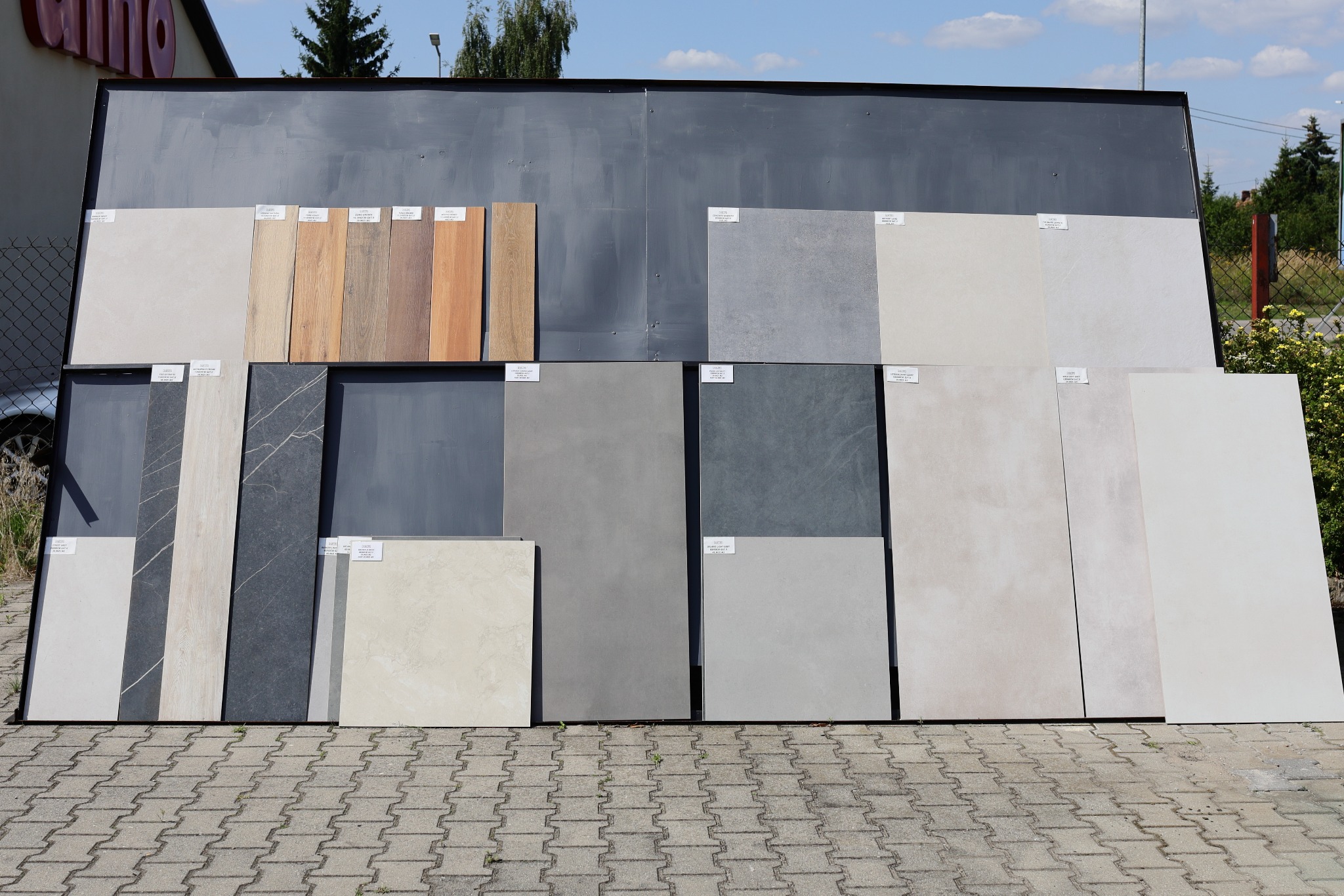 Ekspozycja różnorodnych płytek podłogowych i ściennych w różnych kolorach i wzorach, prezentowanych na stojaku na zewnątrz składu budowlanego.